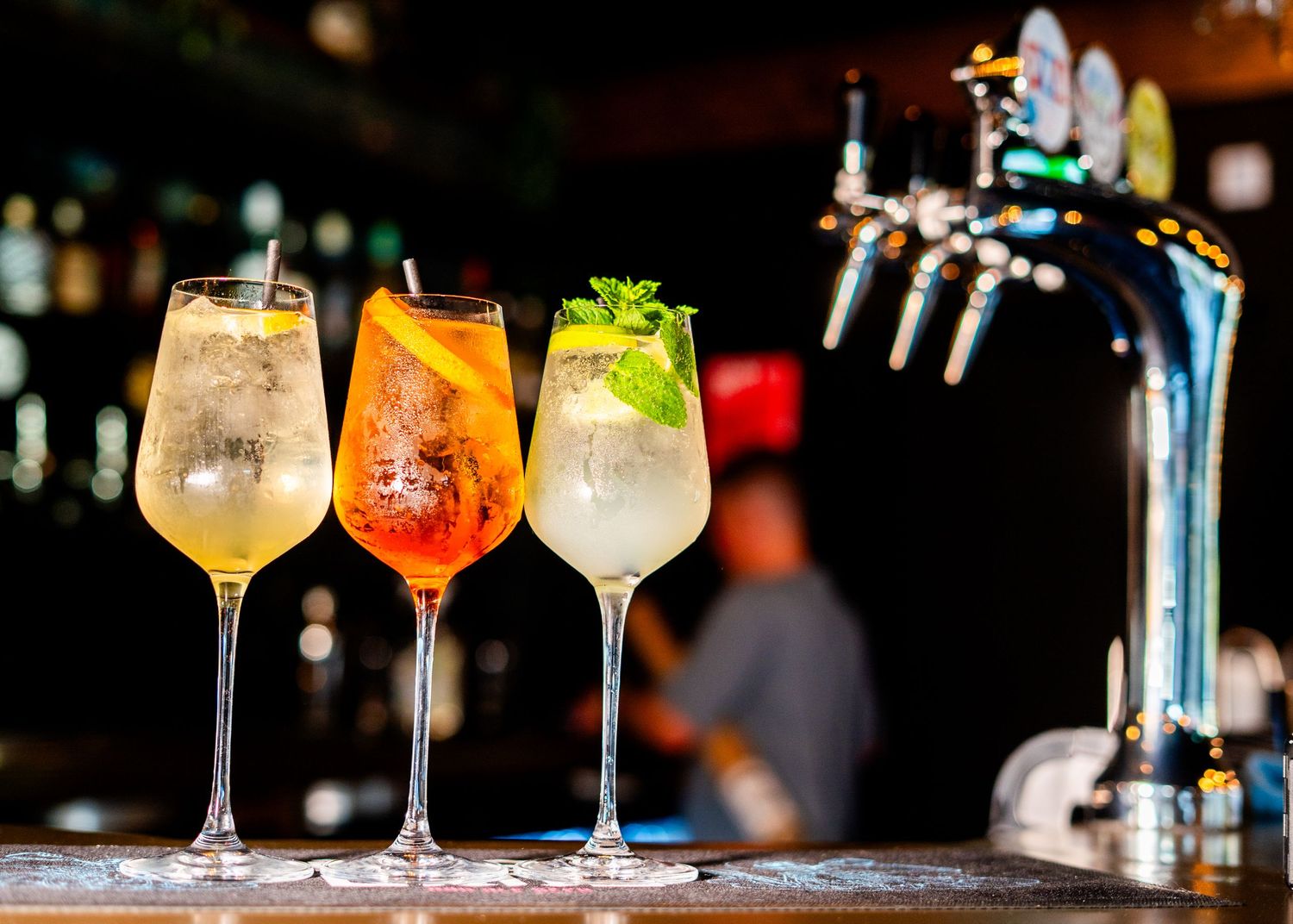Colorful cocktails lined up on a bar counter with garnishes and glassware in elegant lighting.
