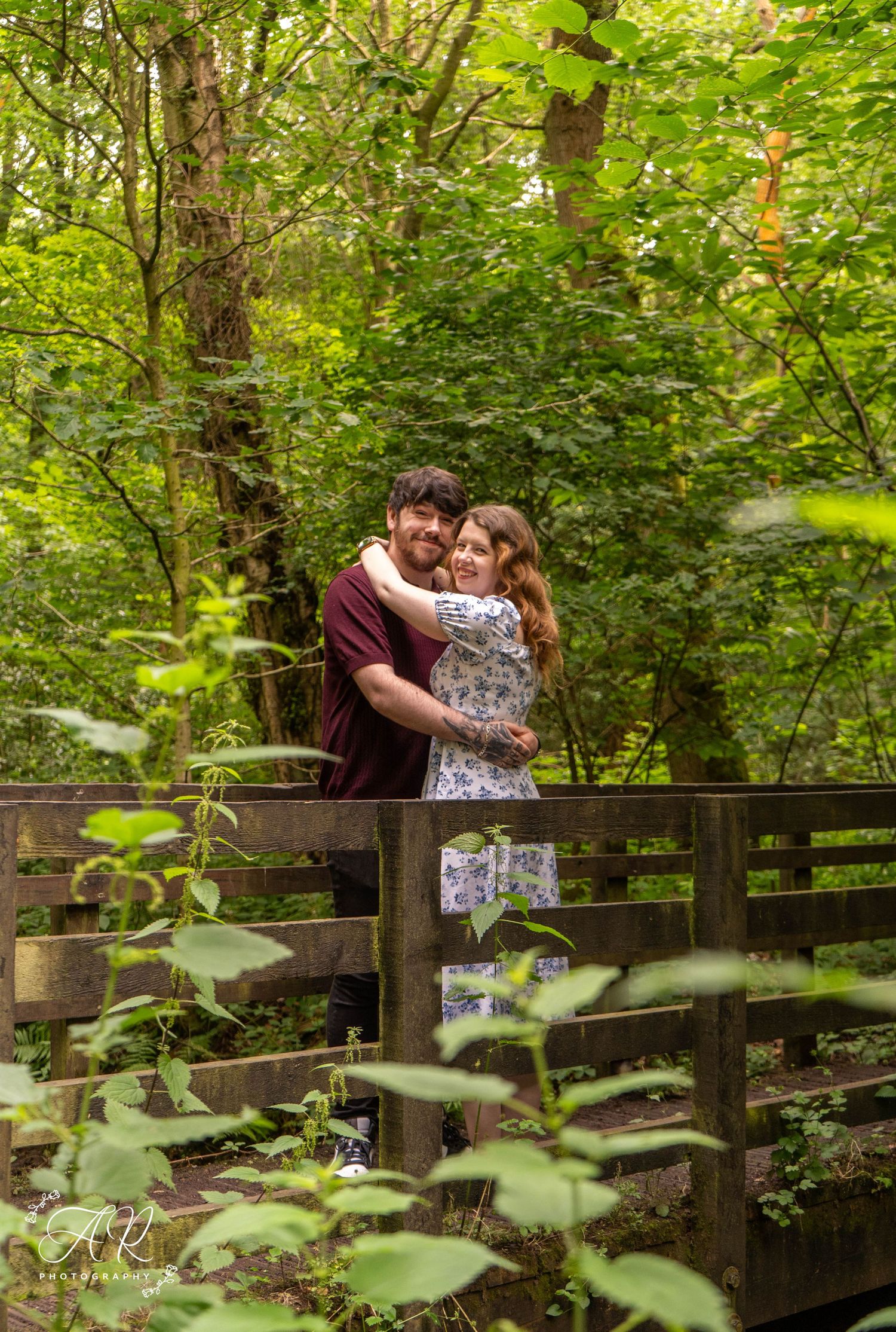 A couple shares an embrace on a wooden bridge surrounded by dense forest foliage.