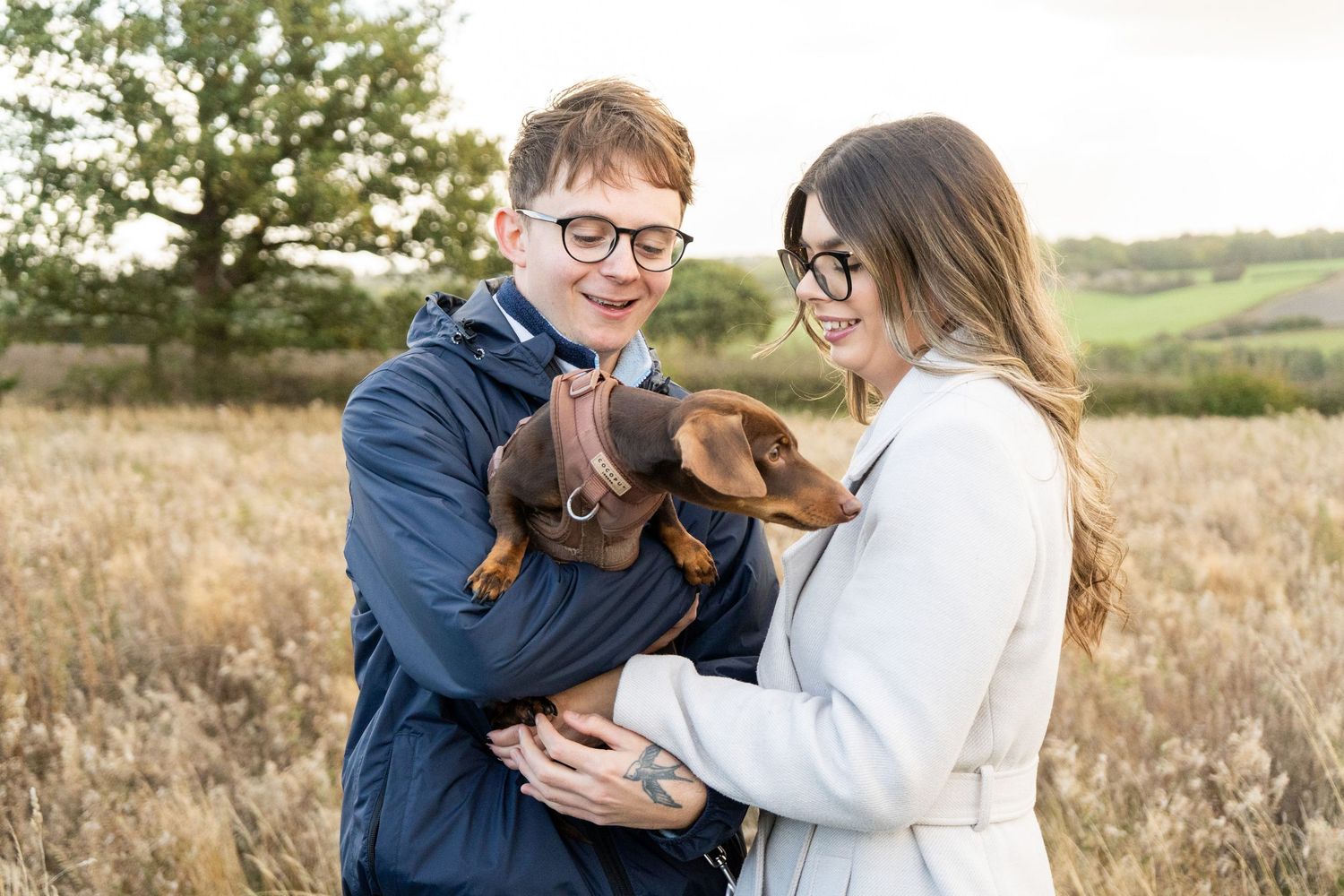 A couple holding their chocolate dachshund puppy in a field during golden hour.