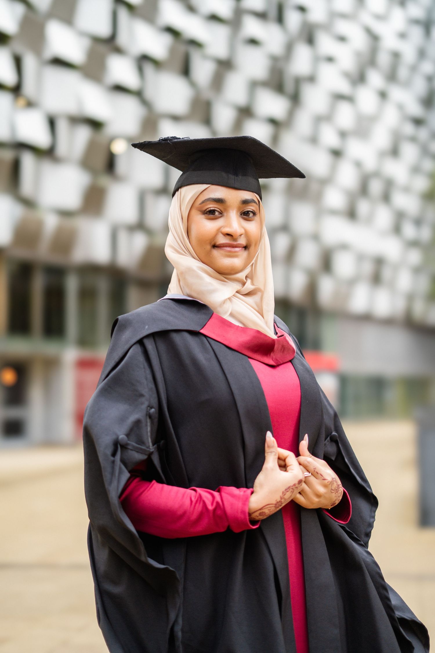 Graduate in traditional academic regalia poses against geometric white wall background.