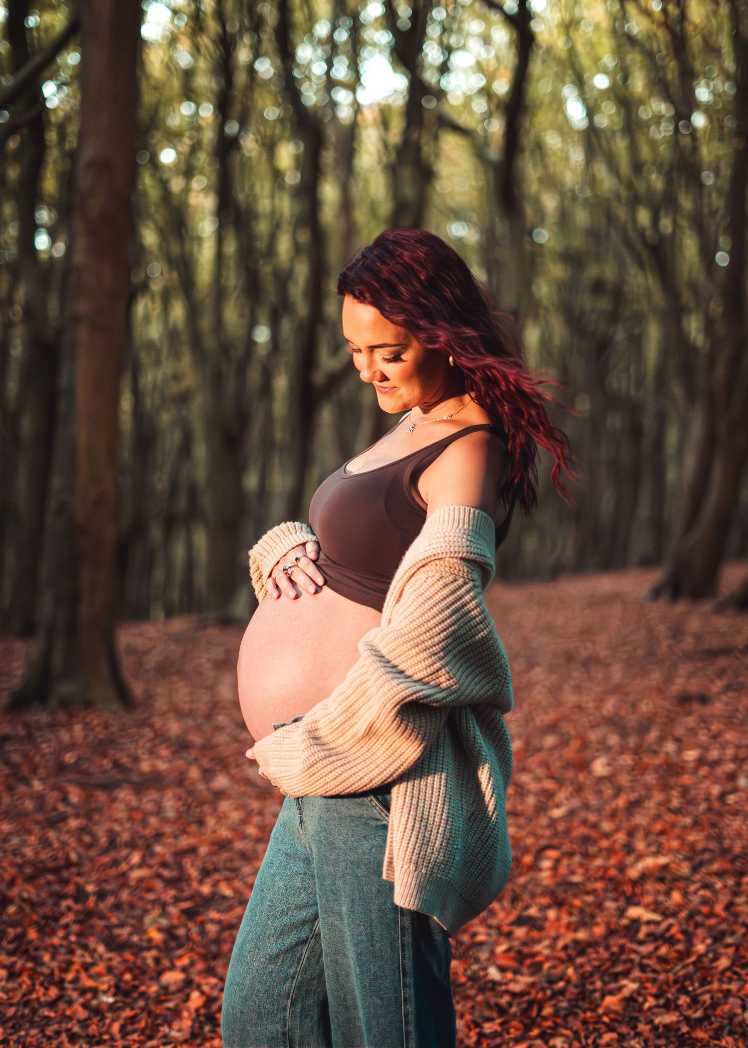Person in cream sweater and jeans poses in autumn forest with fallen orange leaves.
