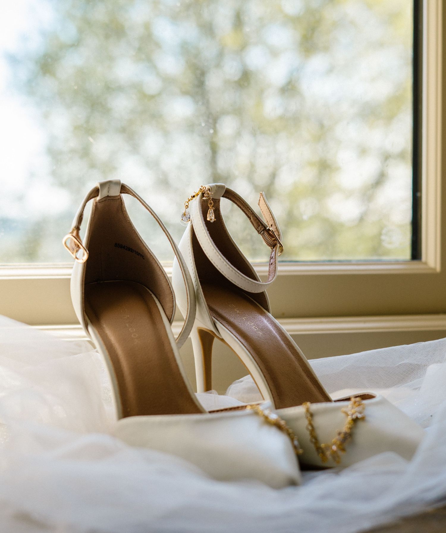 Elegant white bridal shoes displayed on soft white bedding near a window.