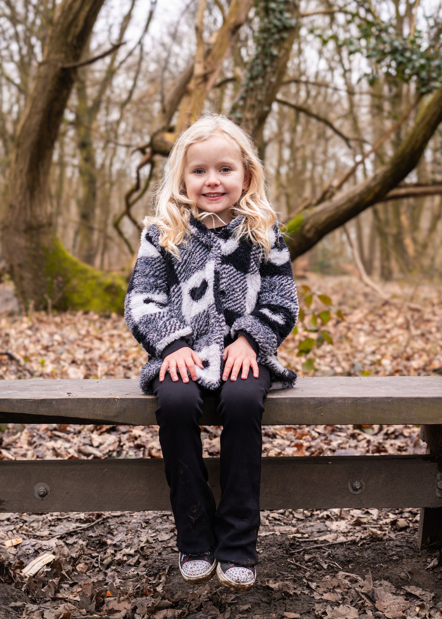 A child in winter clothing sits on a wooden bench surrounded by fallen leaves.