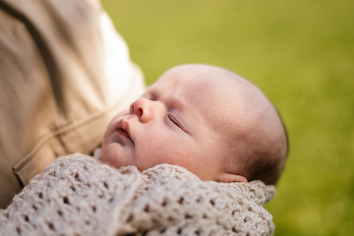 Peaceful sleeping newborn wrapped in cream blanket against green outdoor background.