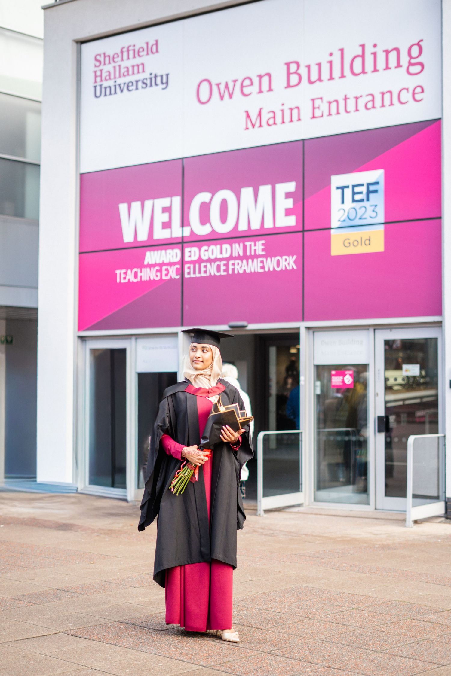 Graduate in academic regalia poses outside Sheffield Hallam University Owen Building entrance.