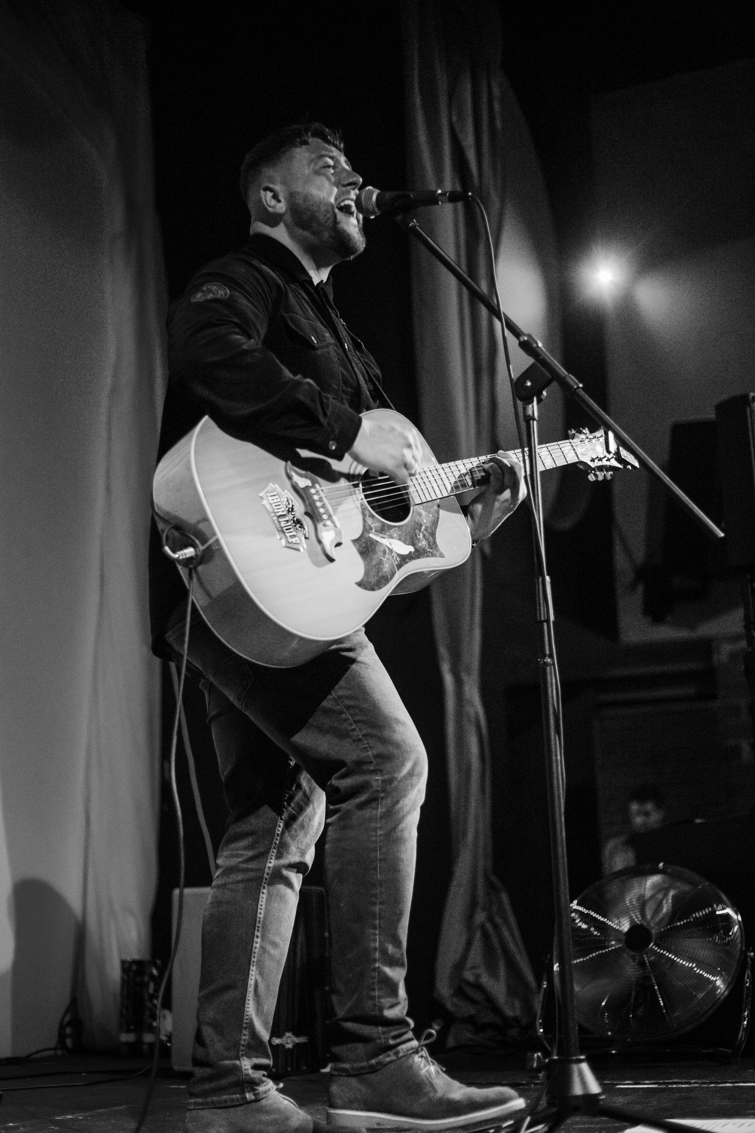 Black and white shot of acoustic guitarist performing passionately on stage with microphone.