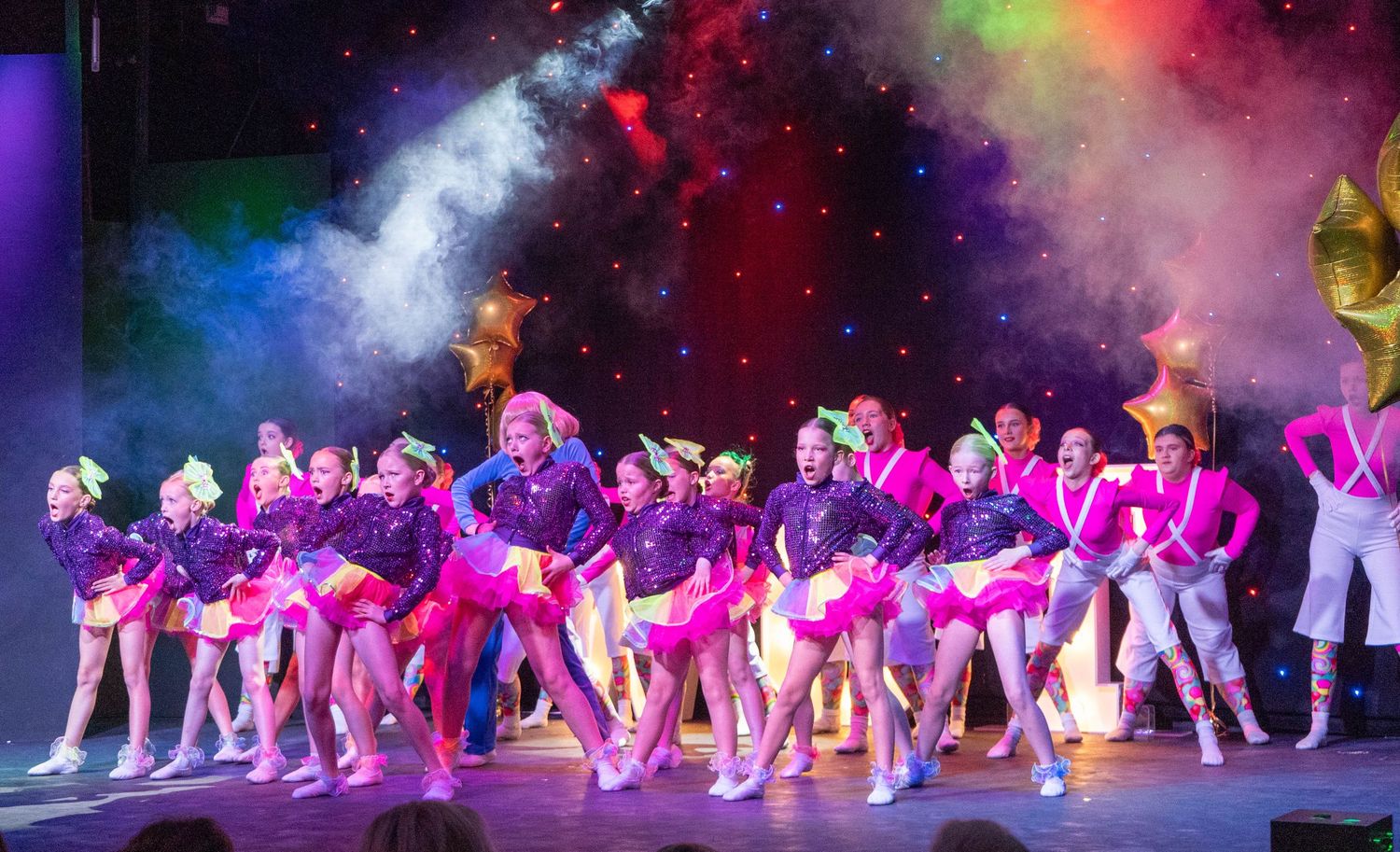 Large group of dancers in matching pink outfits perform under vibrant multicolored stage lights.