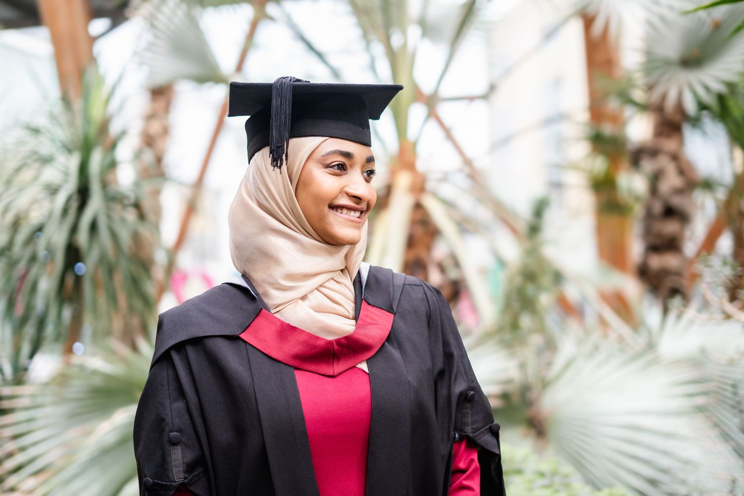 Graduate in black cap and gown poses with loved one in front of palm trees during commencement celebration.