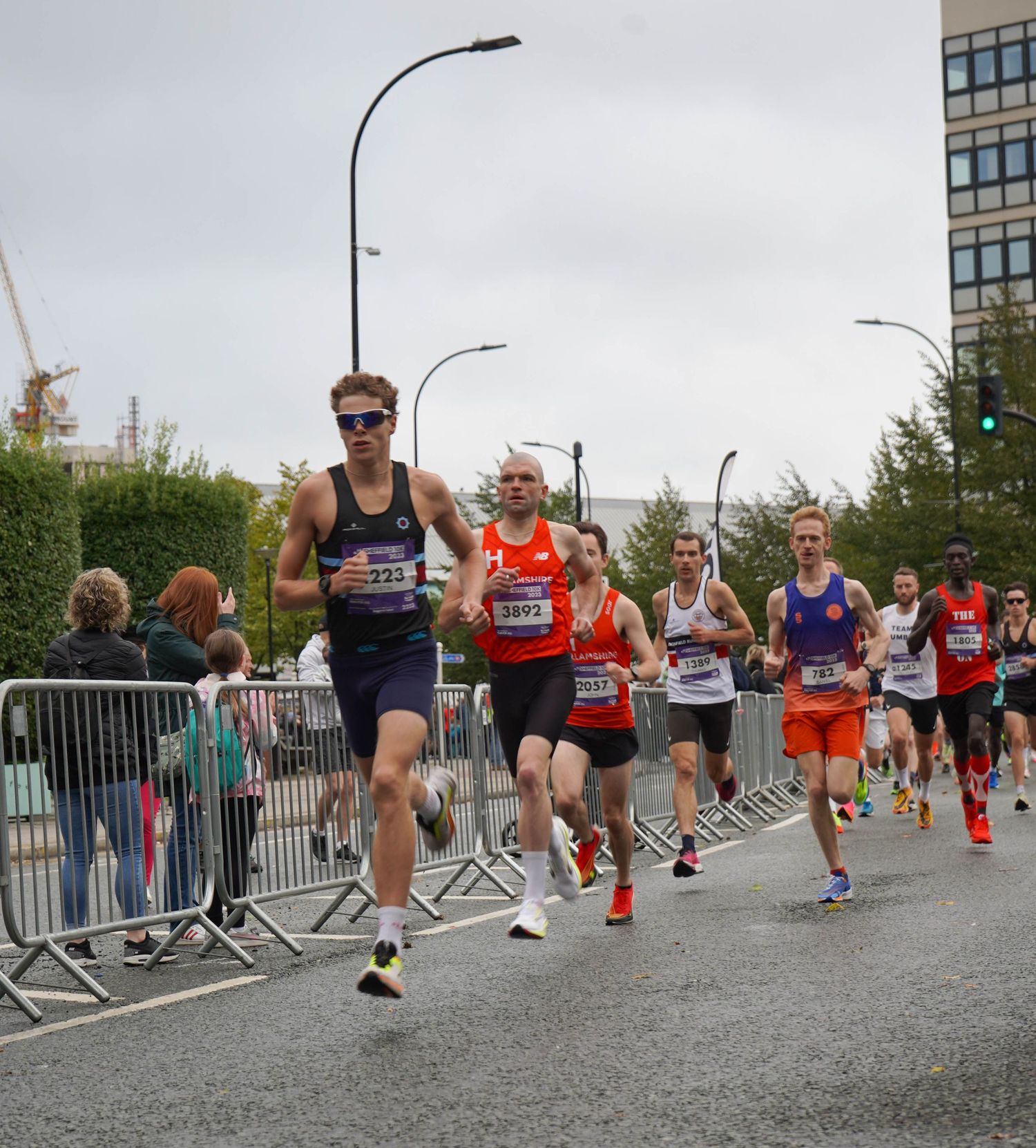 Runners compete in a road race with spectators watching from behind barriers.