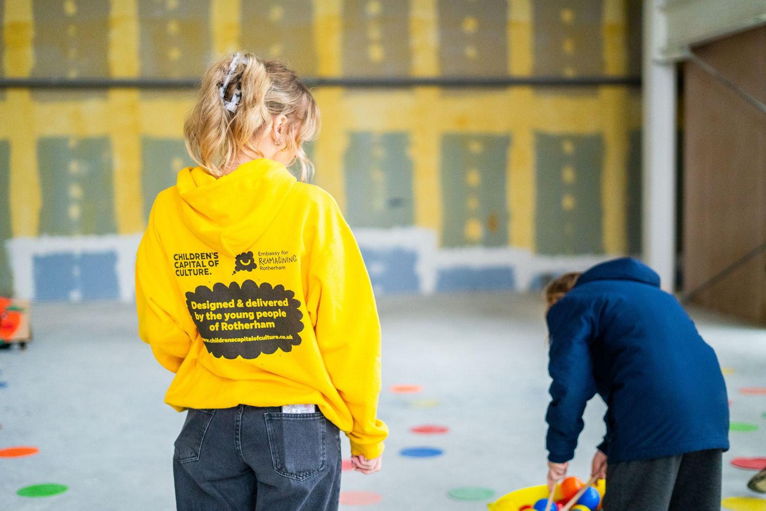 Person in yellow hoodie standing in front of yellow wall with construction materials.