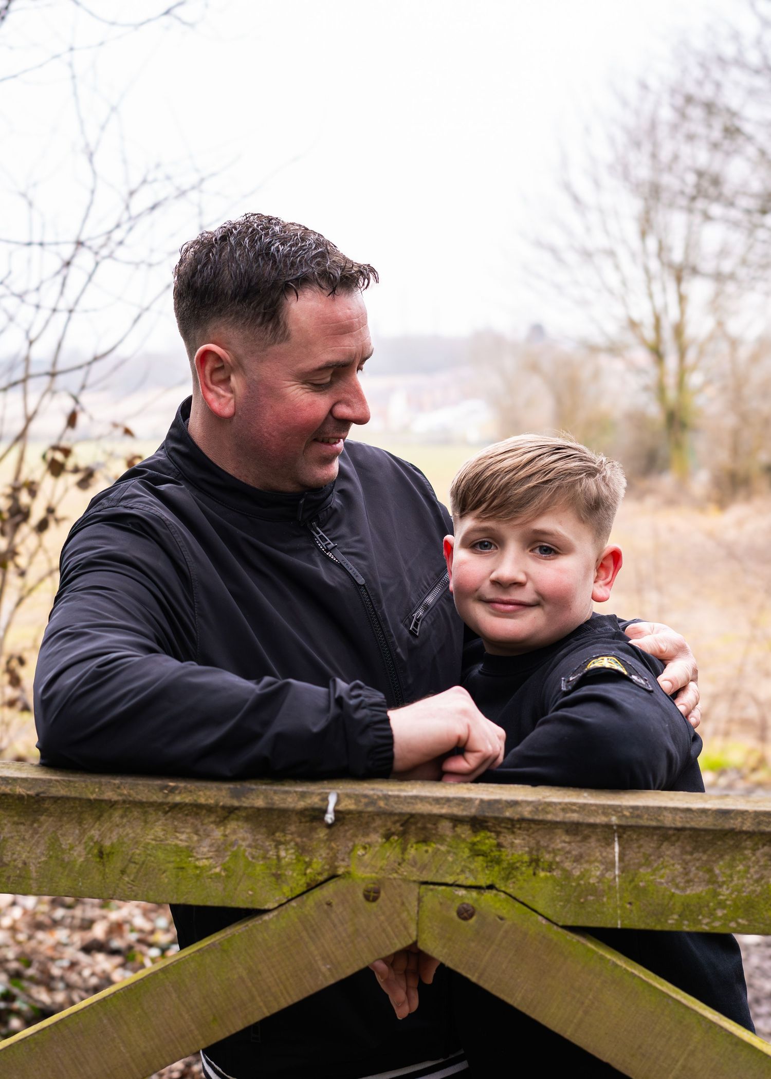 A parent and child share a tender moment while leaning against a wooden gate in nature.