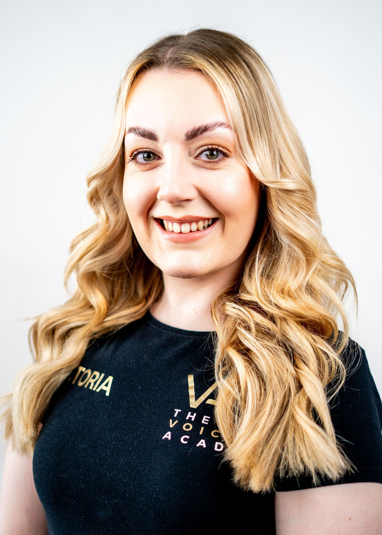 Studio portrait of a person with wavy blonde hair wearing a black uniform shirt with gold lettering.