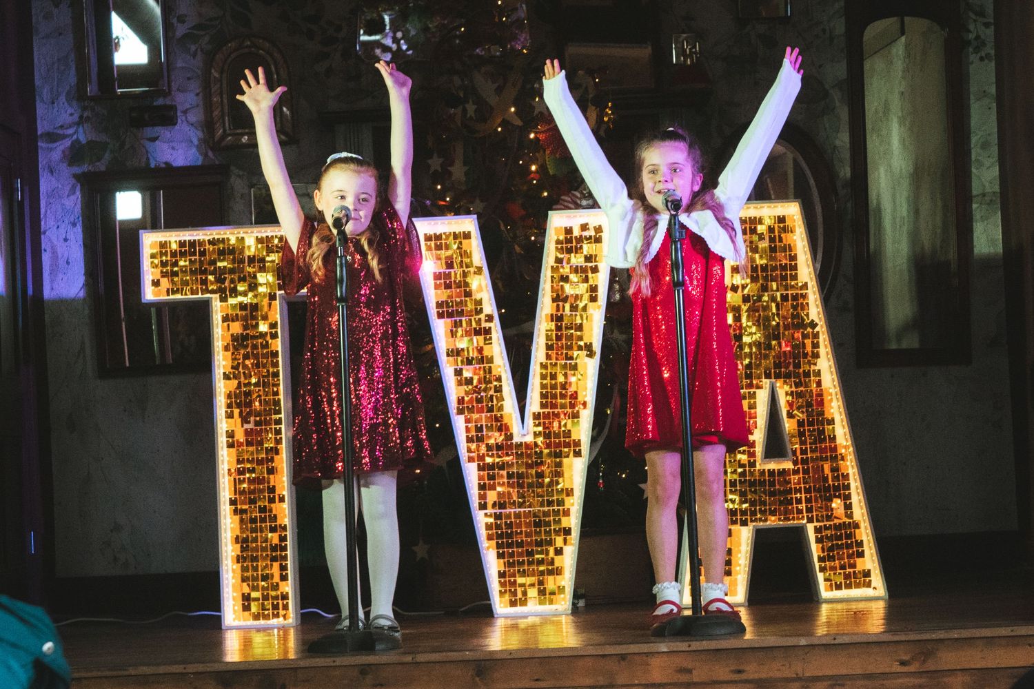 Illuminated letters spell out text against a dark backdrop with performers in red dresses on stage.