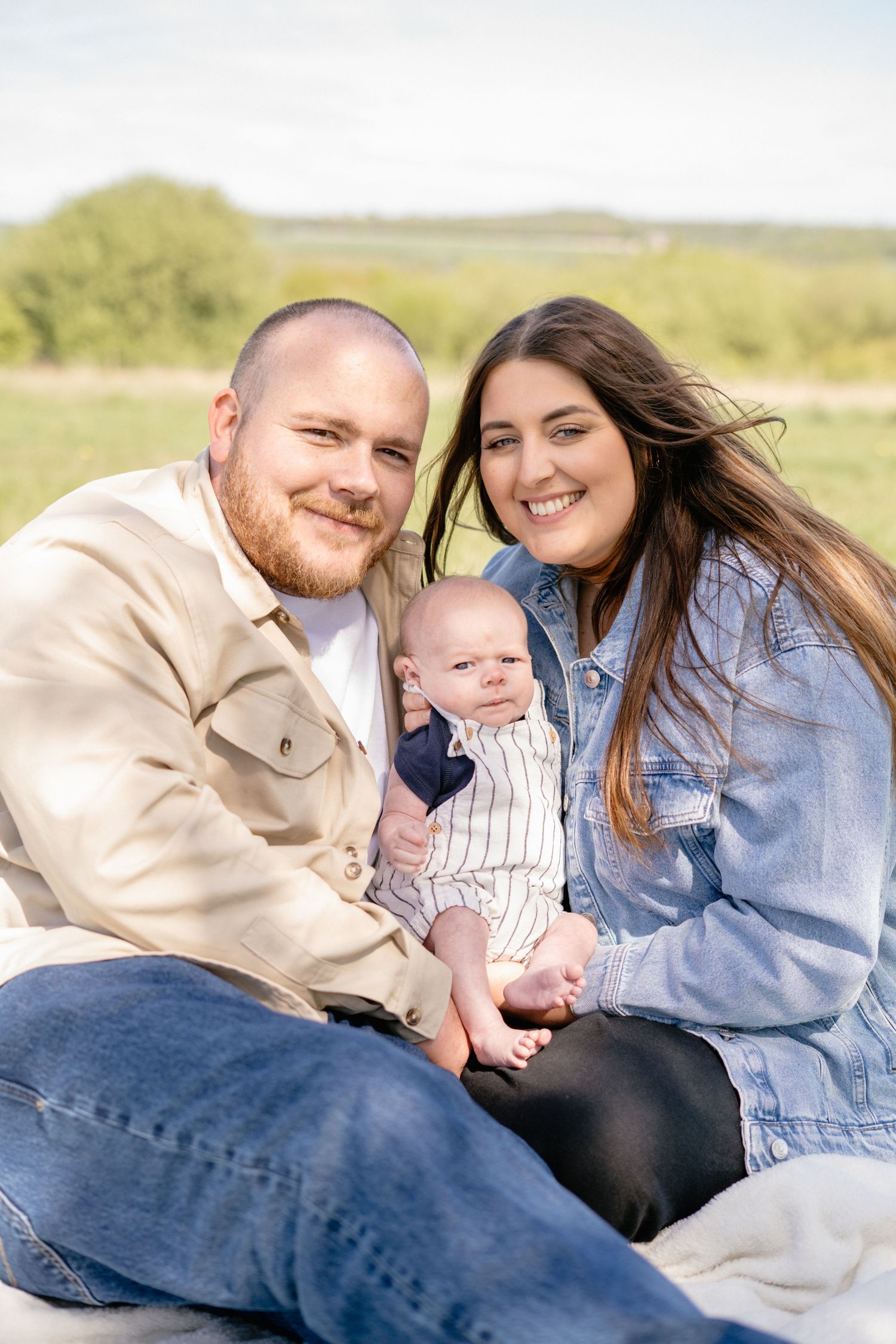 Family smiling and cuddling together on blanket in sunny field.