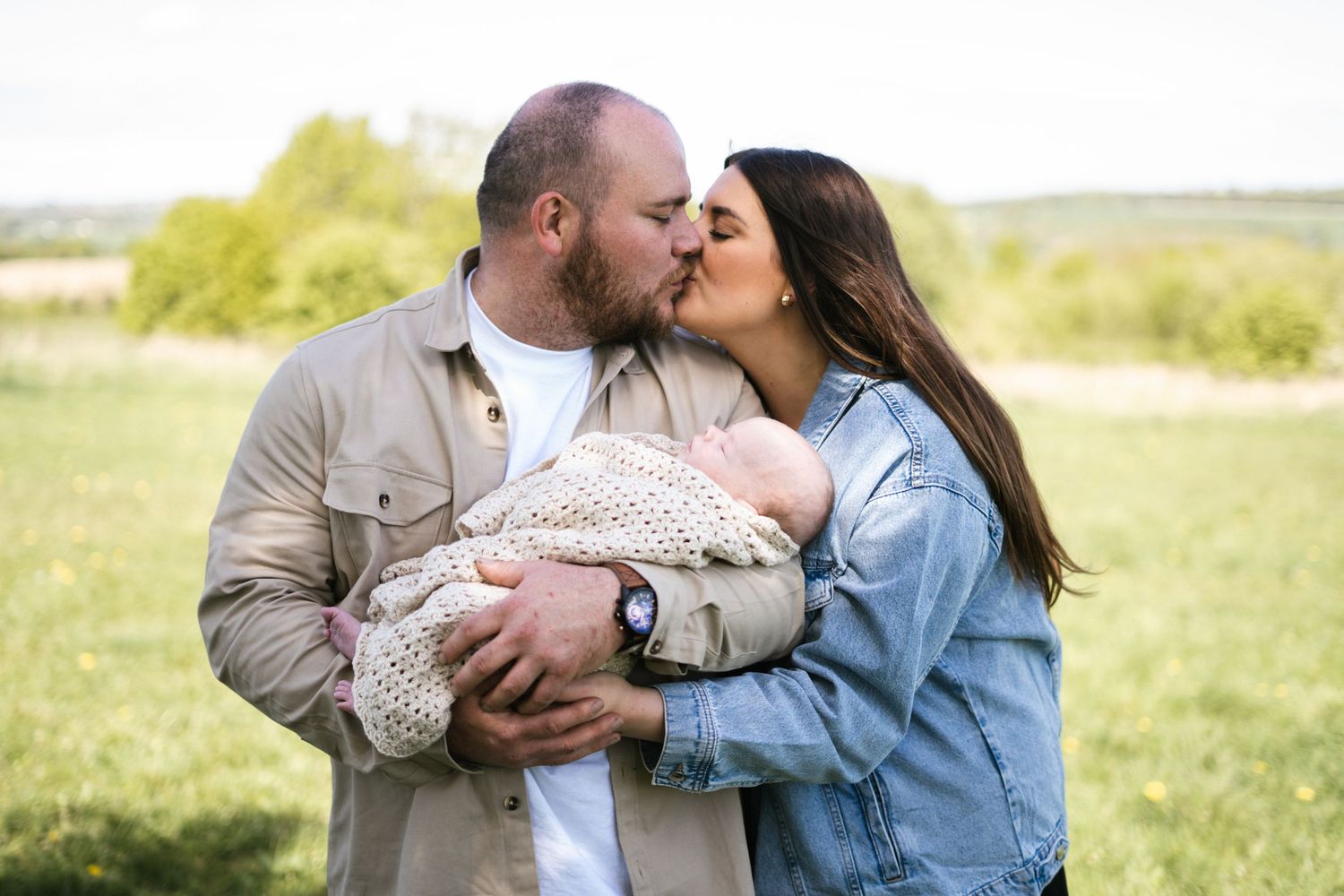 Family embracing tenderly on blanket in sunny meadow with newborn baby.