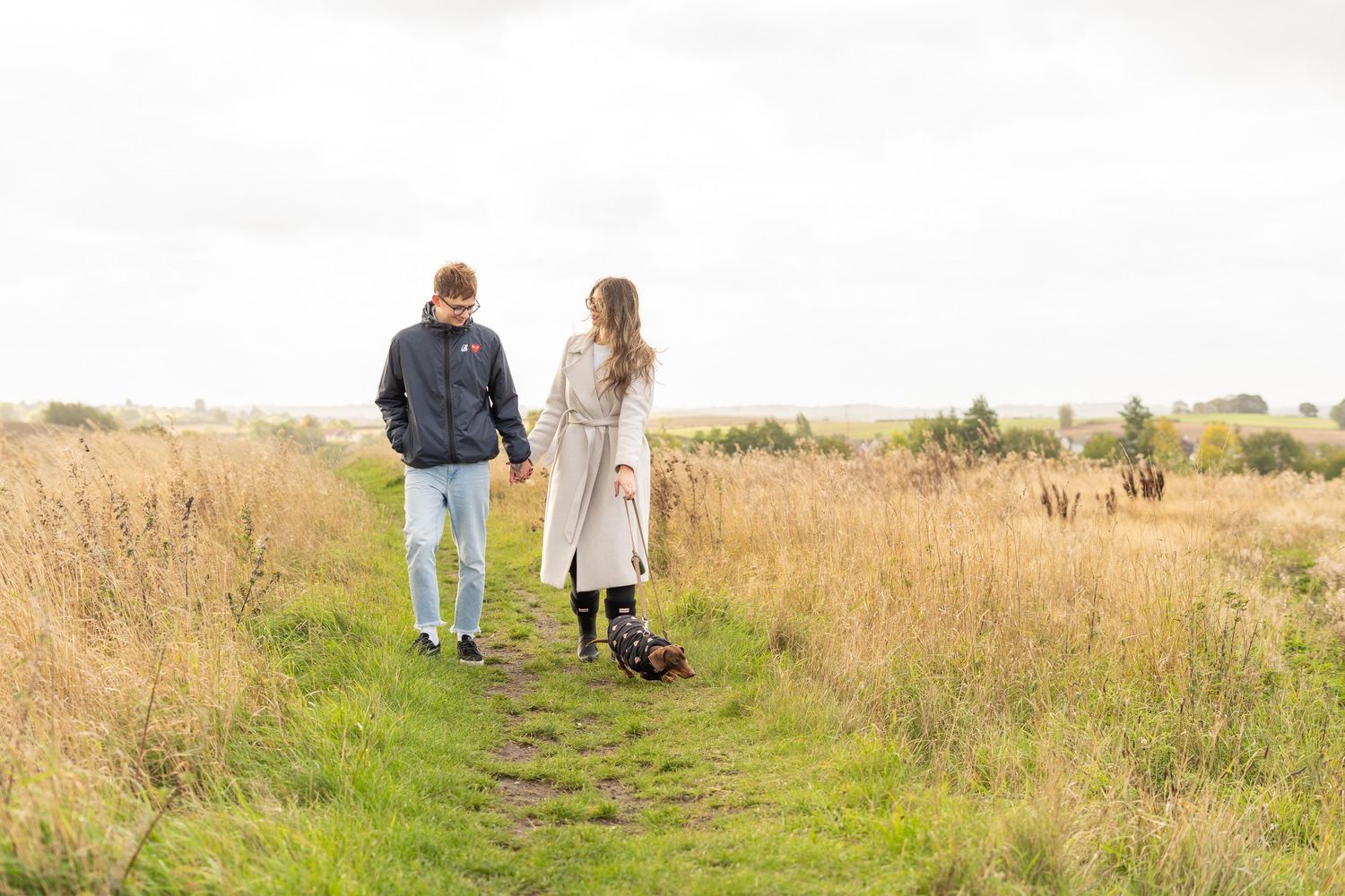 Two people strolling through a scenic meadow with their small dog on a sunny day.