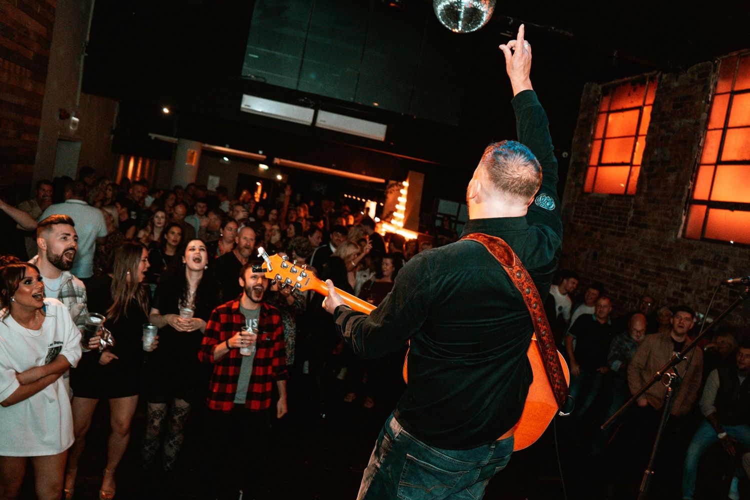 Performer engages crowd under disco ball in dimly lit venue with orange ambient lighting.