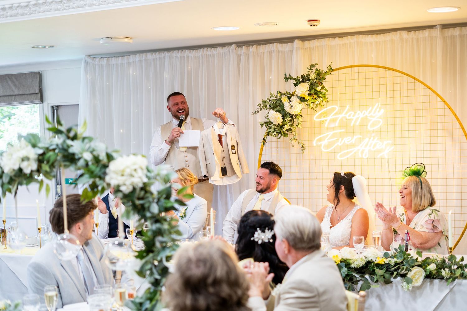 Wedding reception speeches at decorated head table with floral arrangements and neon sign.