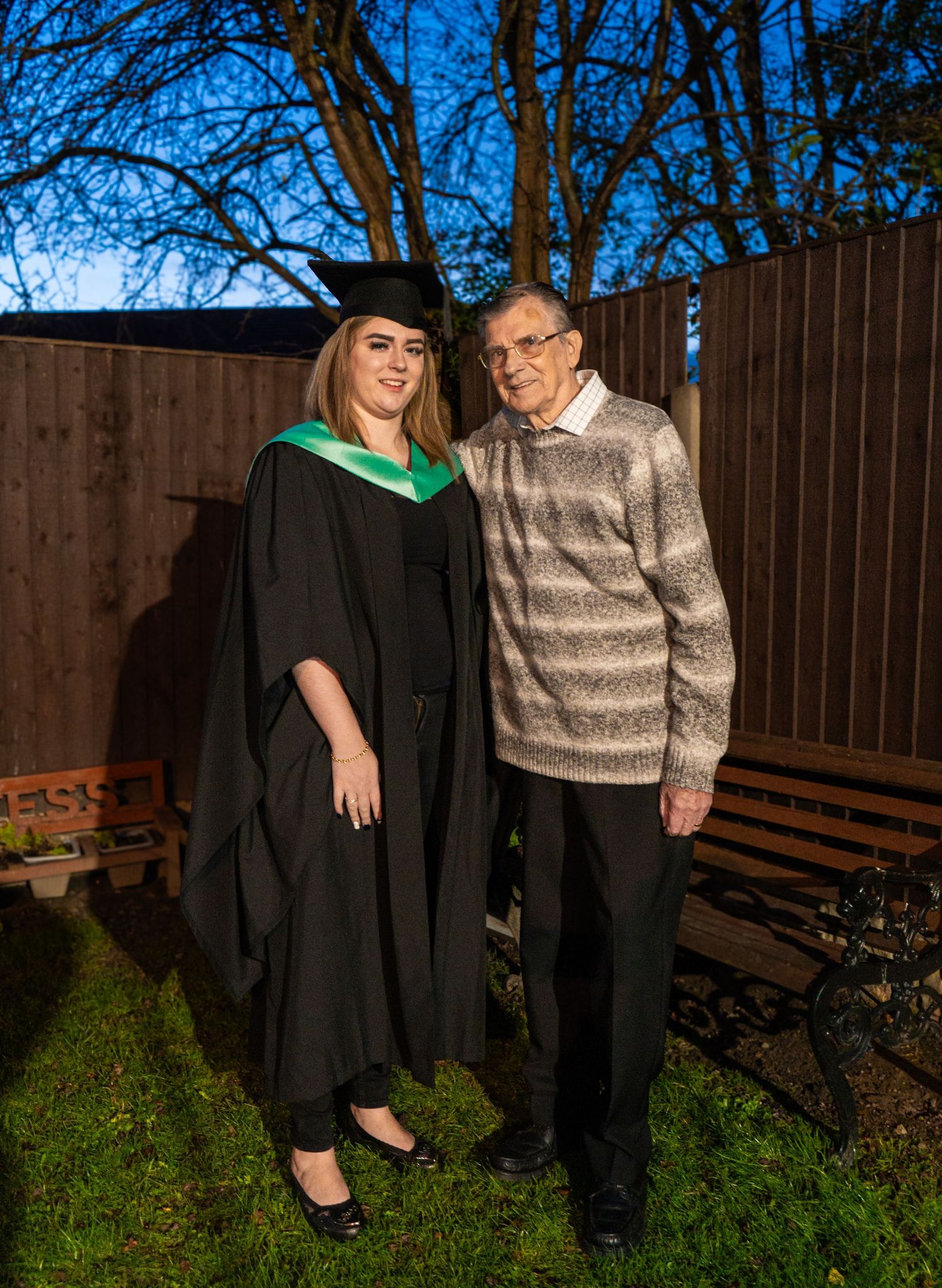 Outdoor graduation photos with graduate and elderly relative against wooden fence background.