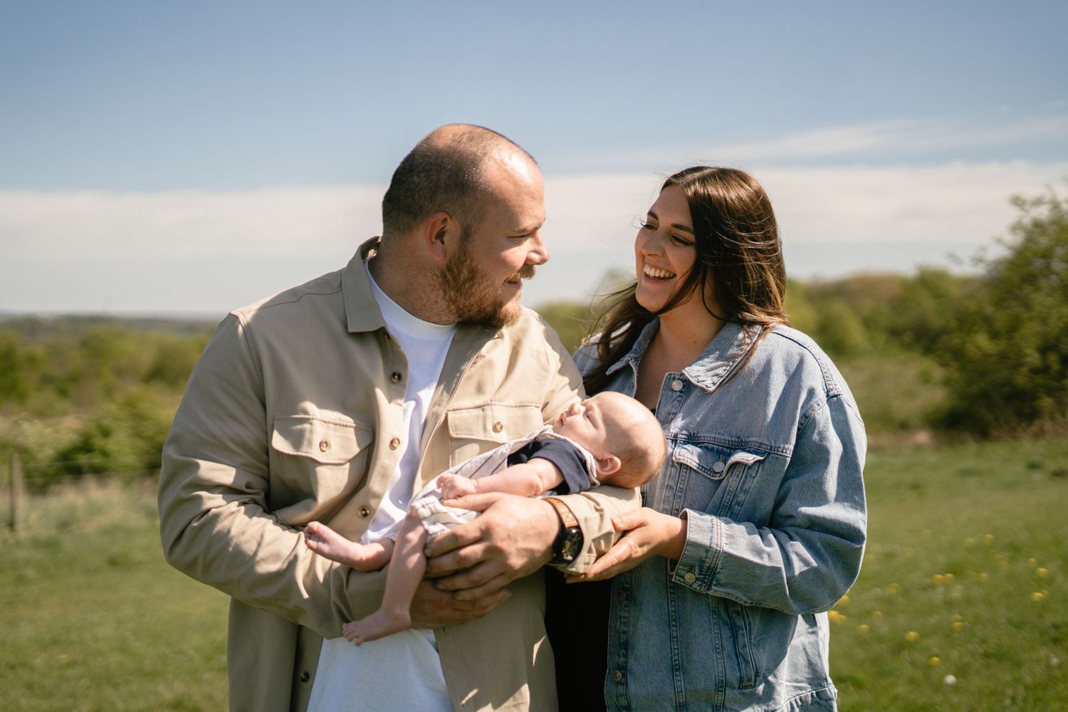 A couple stands in a meadow holding their newborn baby on a bright sunny day.