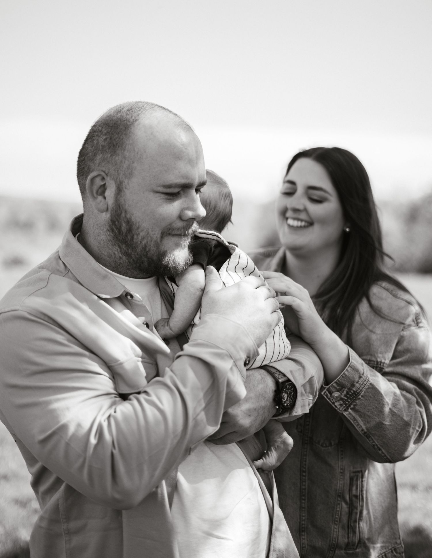 Black and white photo of joyful parents sharing a moment with their newborn outdoors.
