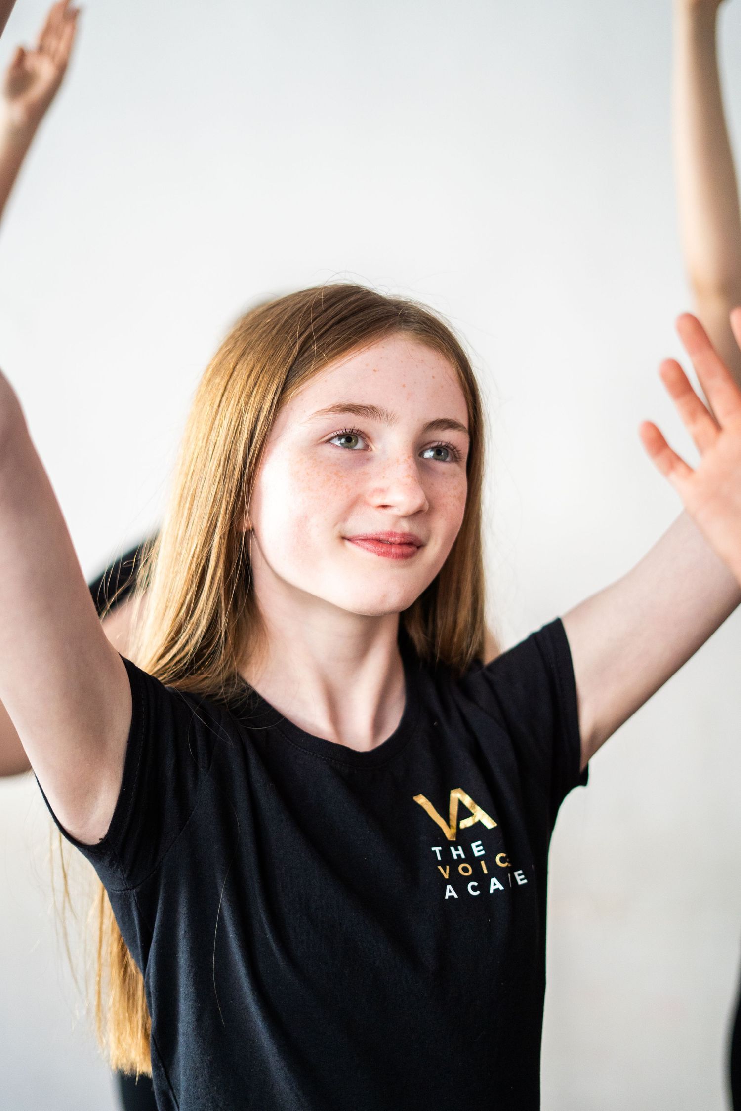 Students in matching black t-shirts practice dance moves with raised arms in a studio setting.