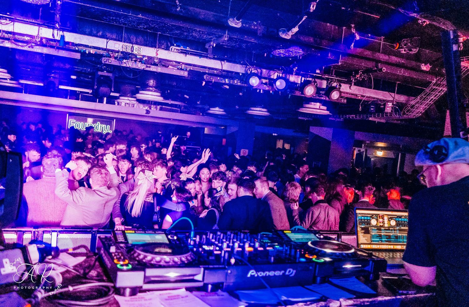 Crowded dance floor with DJ booth and vibrant purple lighting at a nightclub event.