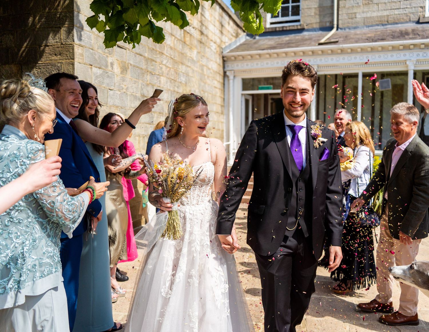 Wedding celebration outside stone building with guests throwing confetti.