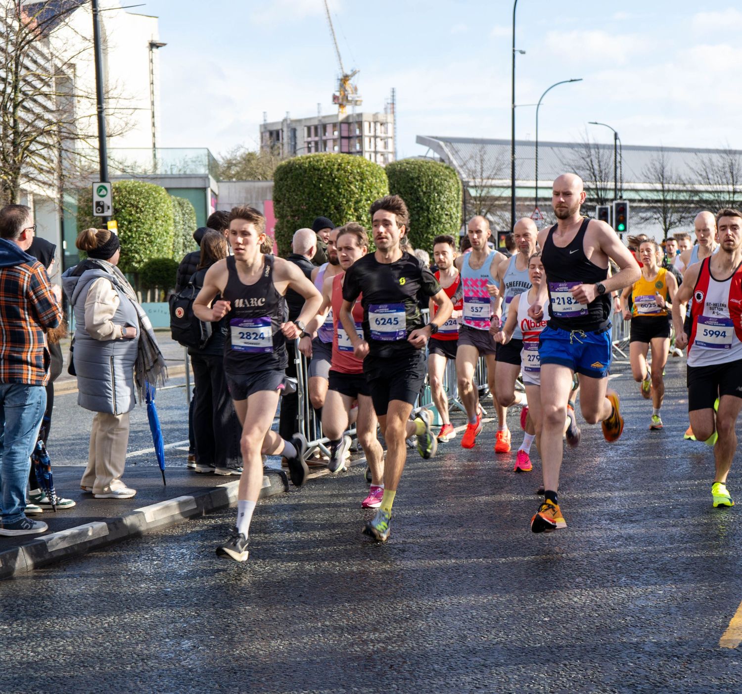 Group of runners compete in city marathon along wet streets with spectators cheering.
