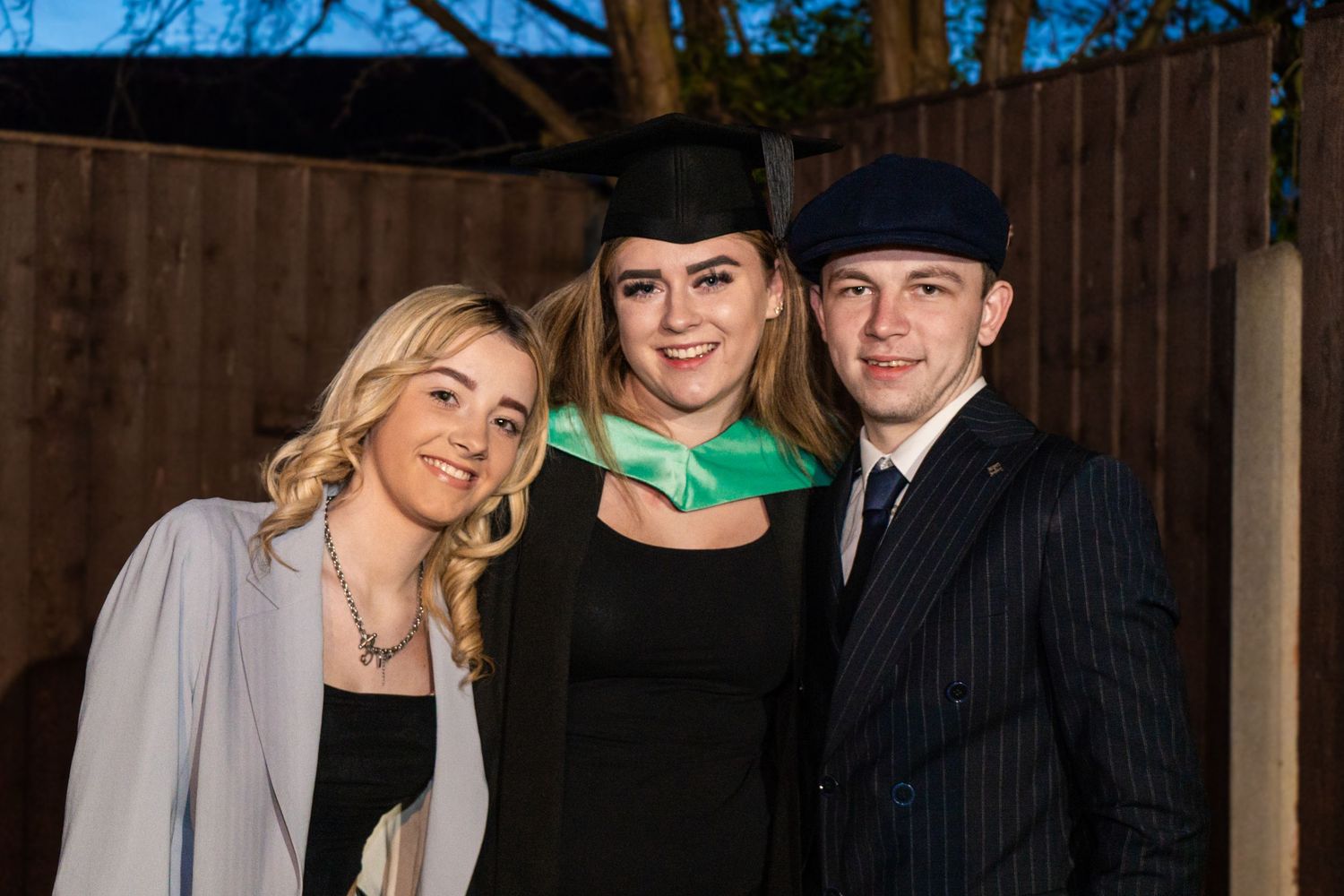 Outdoor graduation portraits with family members against wooden fence and blue sky.