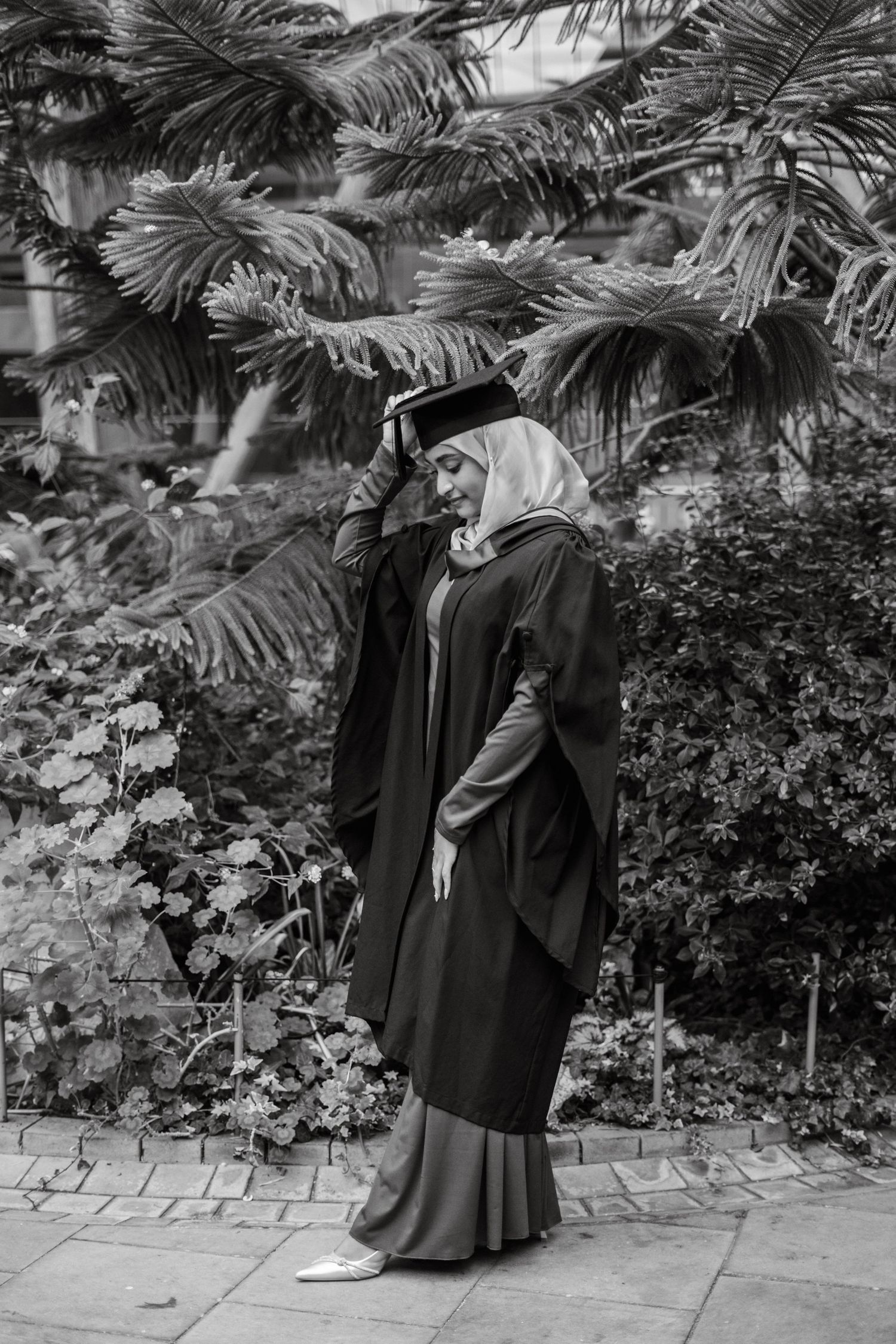 Graduate in black cap and gown poses against tropical foliage backdrop during graduation photoshoot.