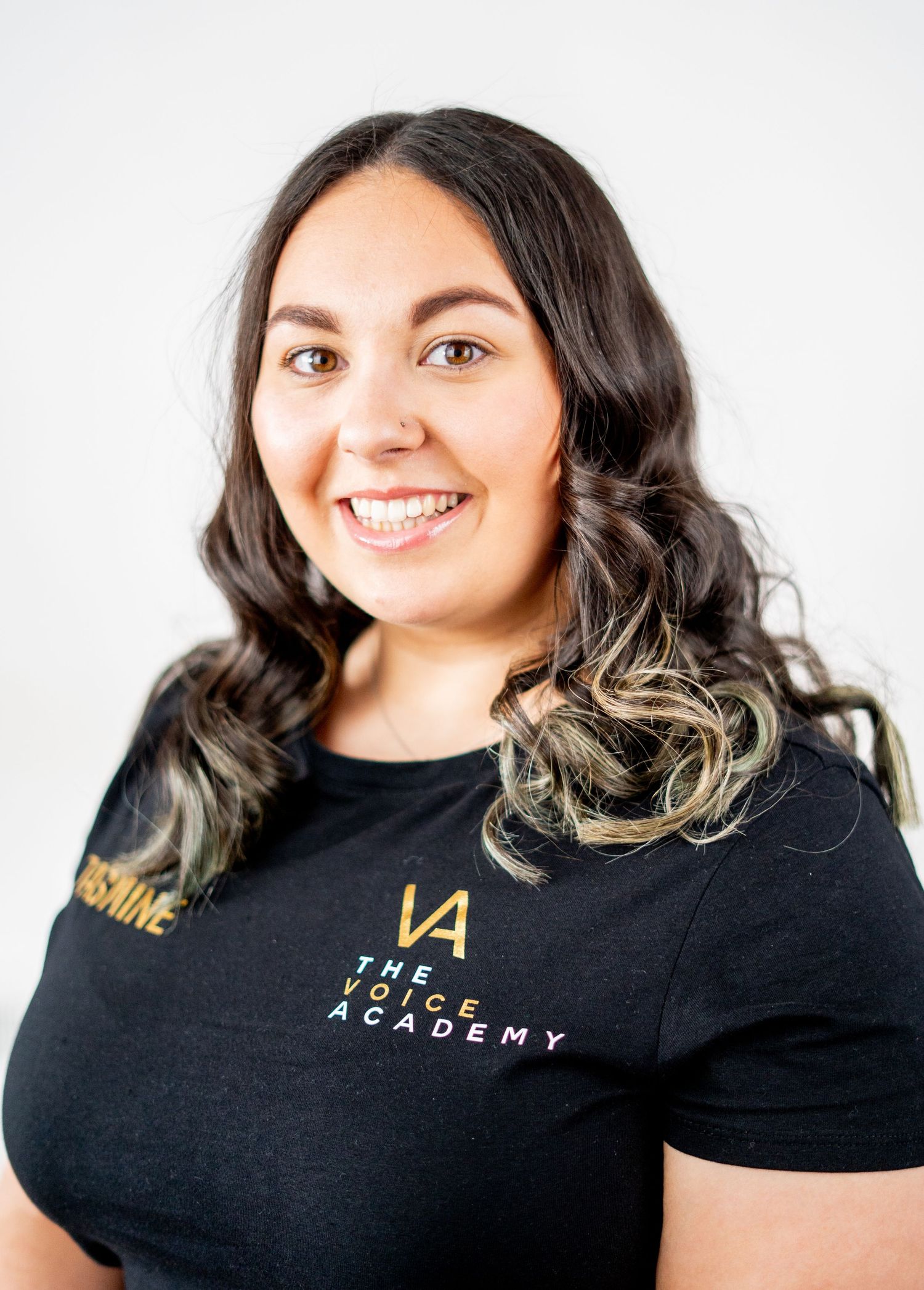 Studio portrait of a person with dark wavy hair wearing a black branded t-shirt.