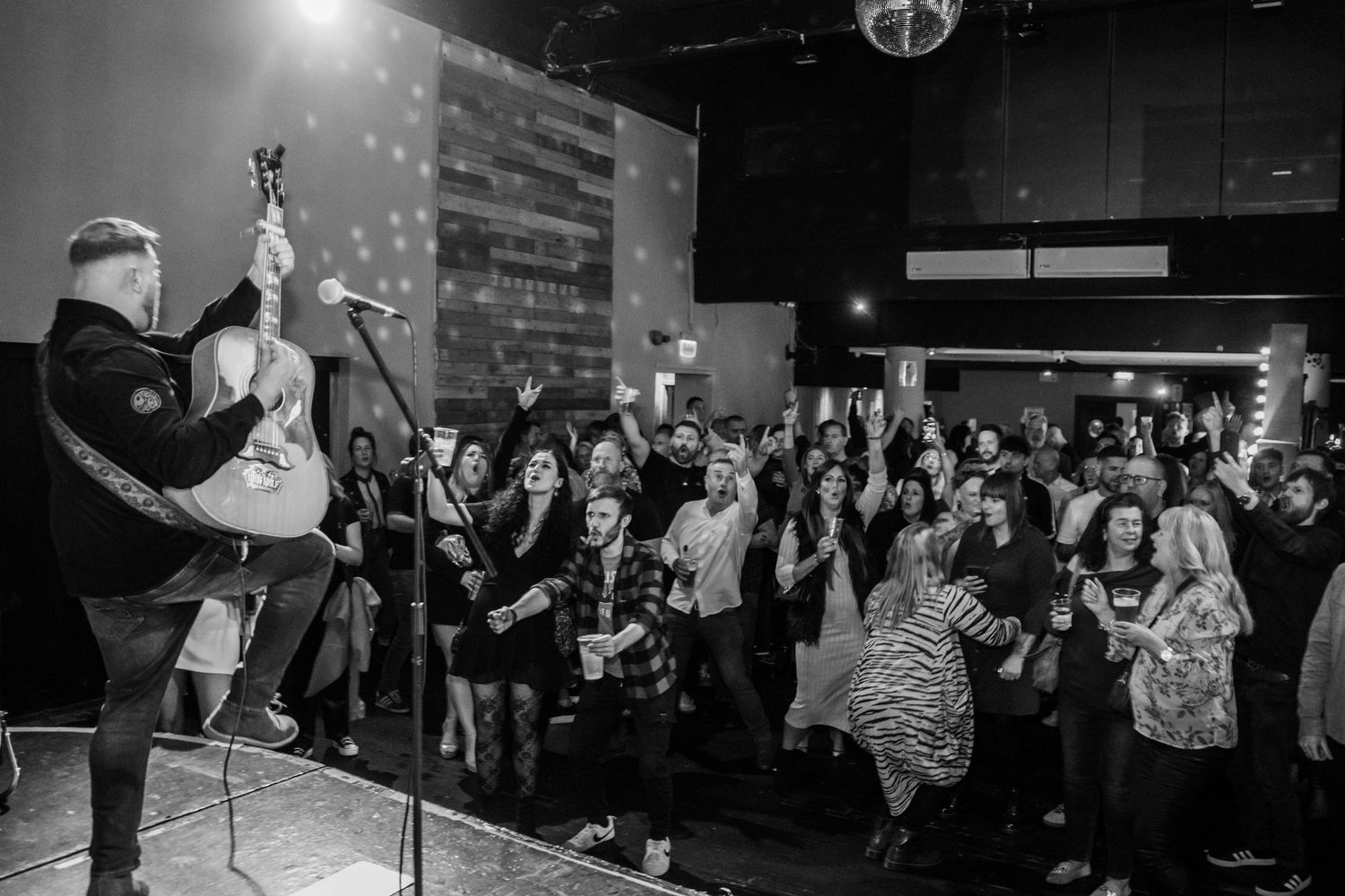 Black and white concert photo showing energetic crowd with raised hands watching performer on stage.
