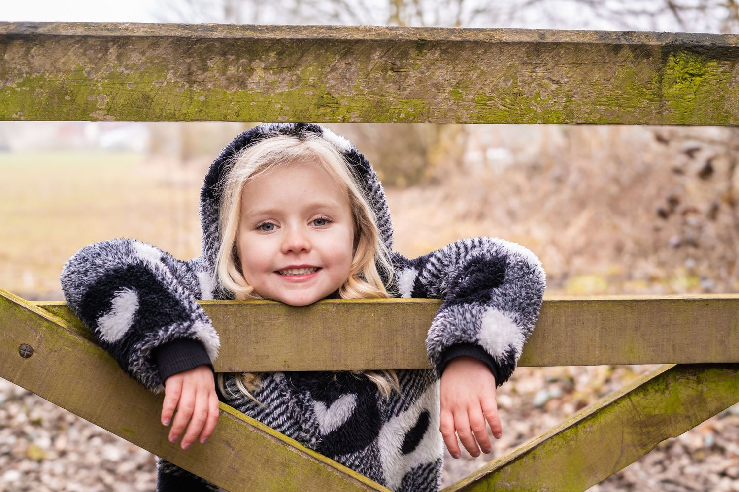 A cheerful person in a winter coat poses playfully between wooden fence slats.