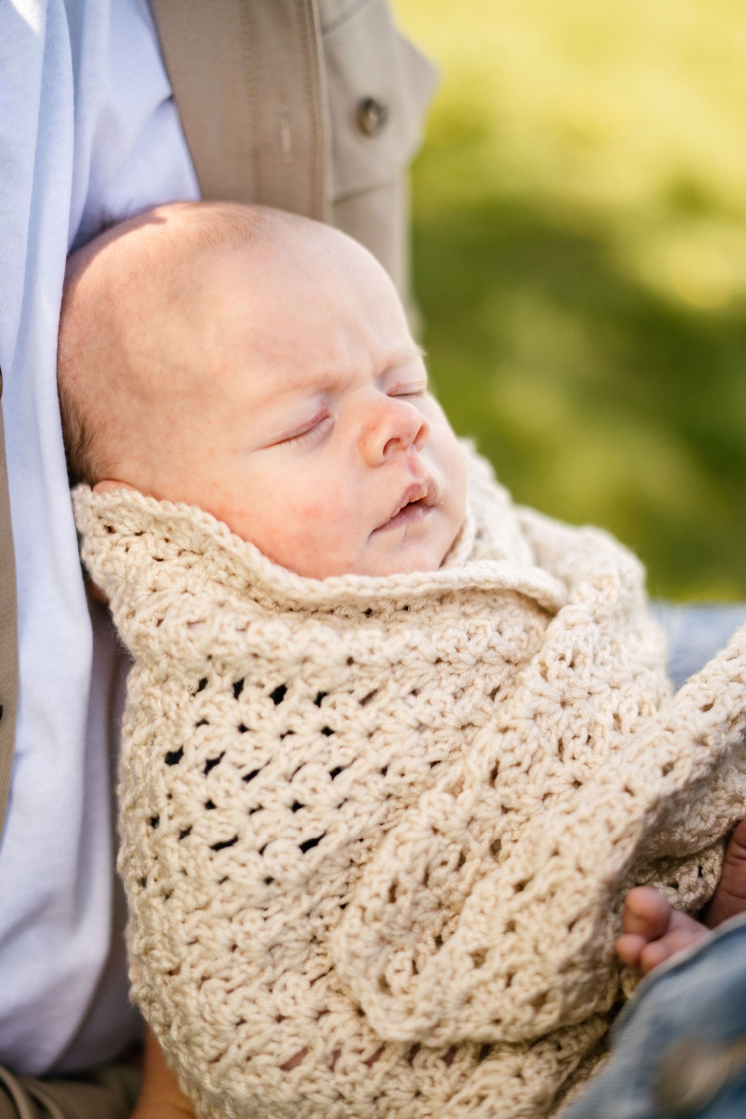Close-up of sleeping infant swaddled in cream crochet blanket against blurred natural background.