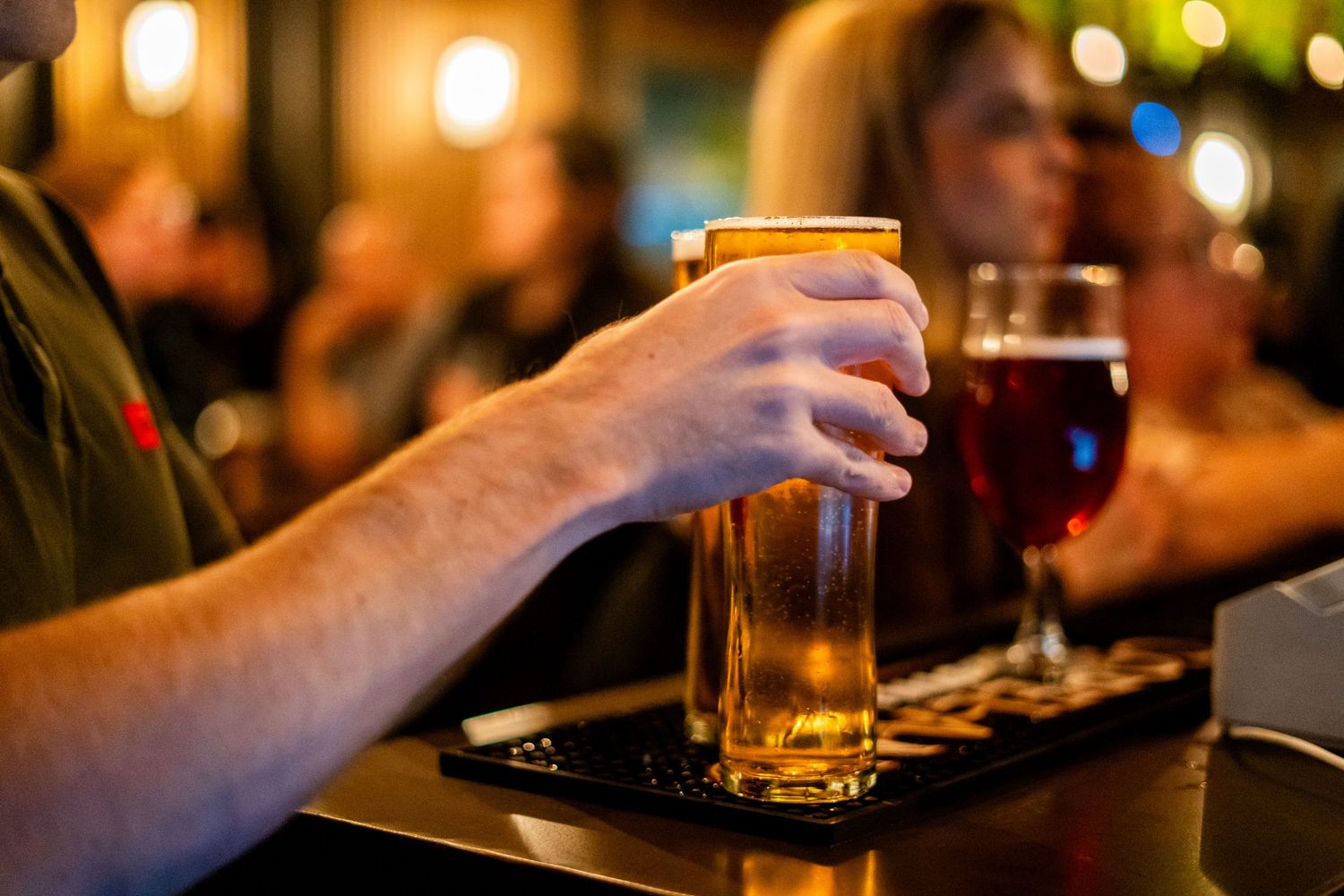 Hand reaching for glass of beer on dark bar counter with blurred background.