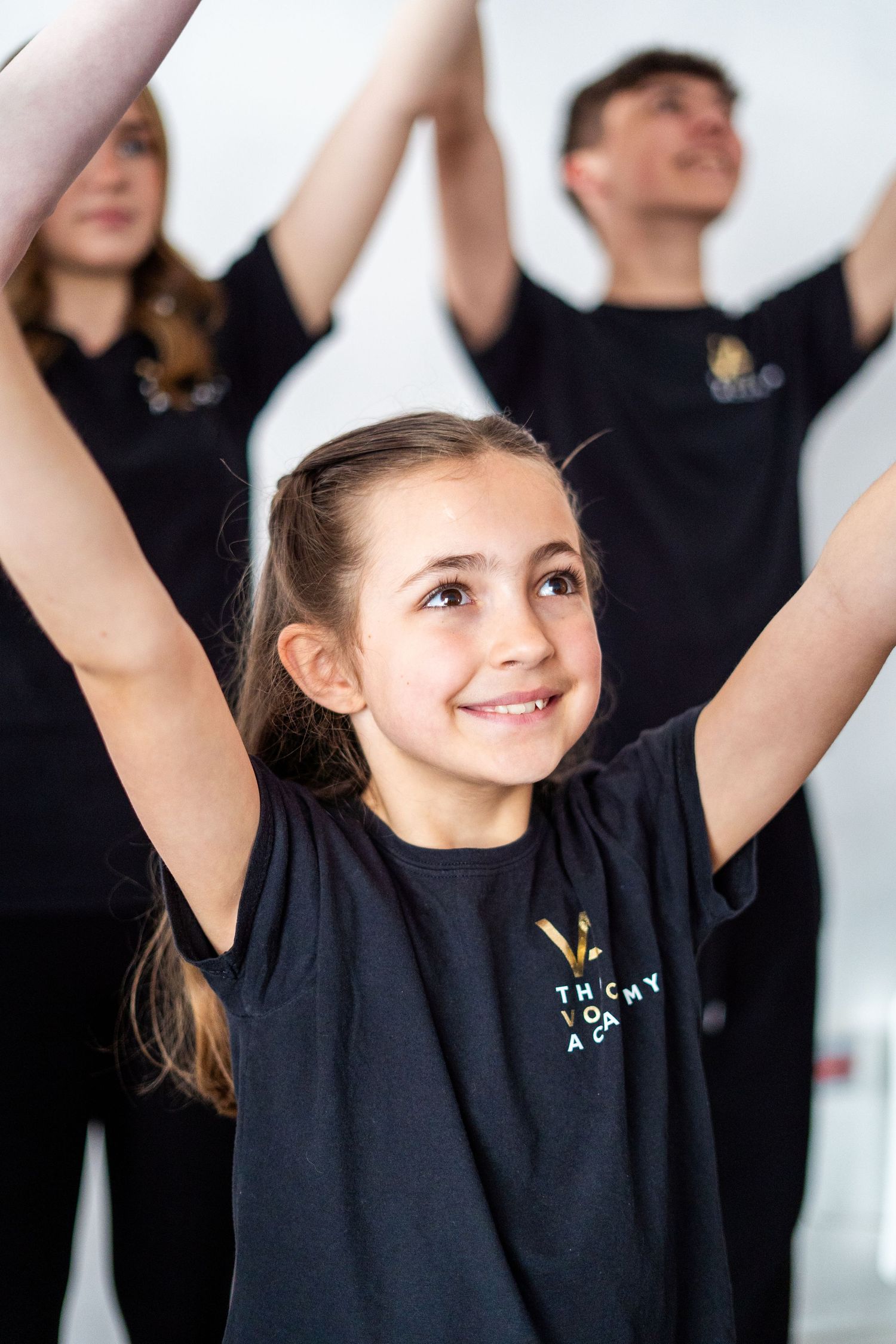 Young dancers in black t-shirts raise their arms during dance practice against a white background.