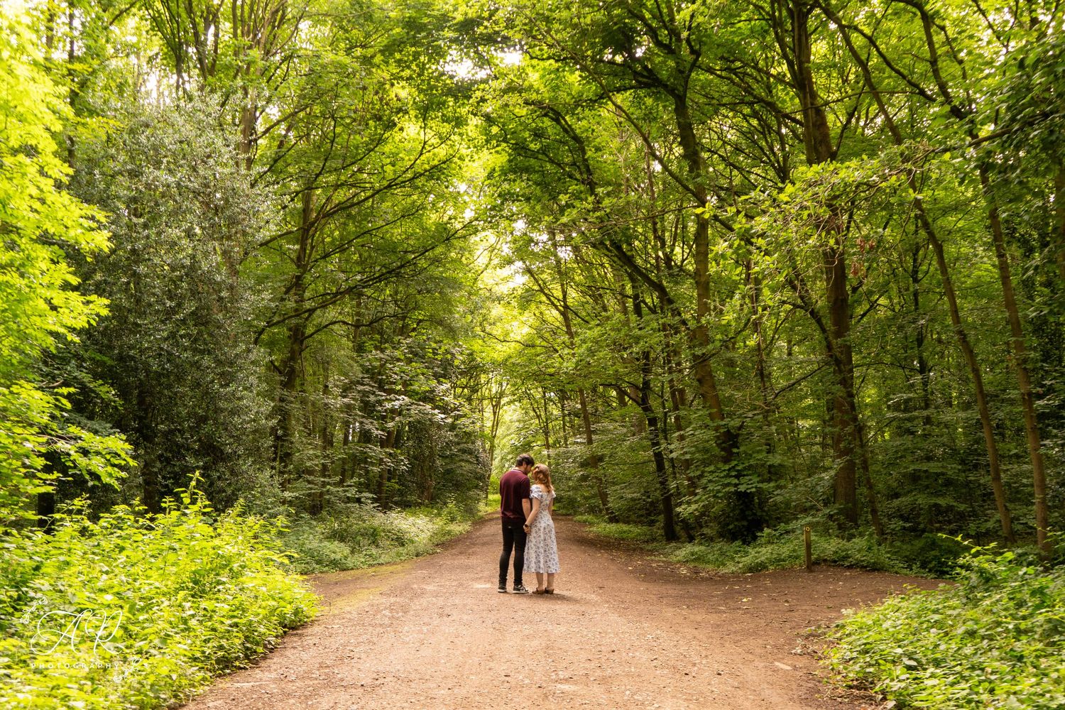 A couple stands together on a sunlit forest path surrounded by vibrant green trees.