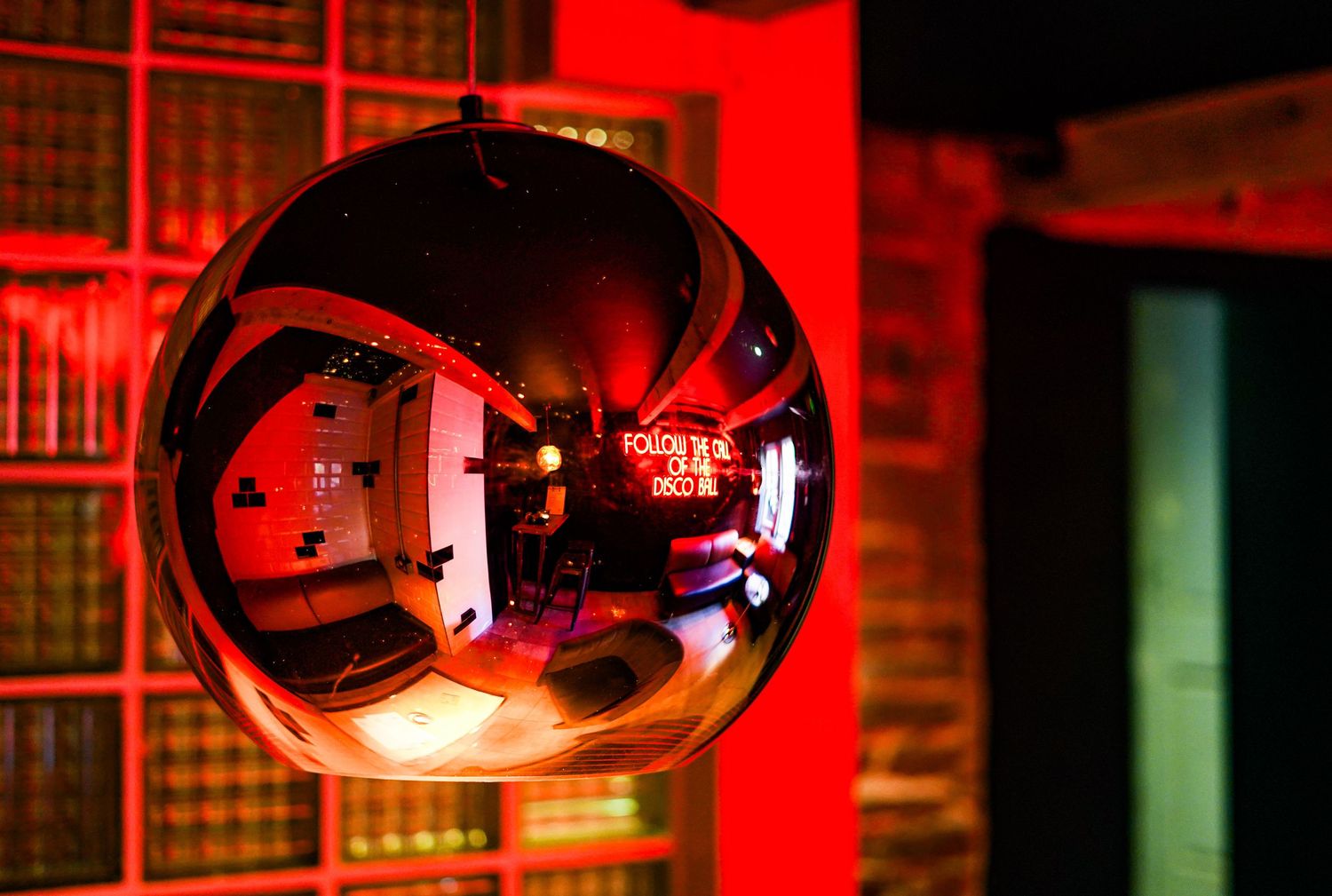 A reflective copper sphere pendant lamp creates a distorted view of a dimly lit room with bookshelves.