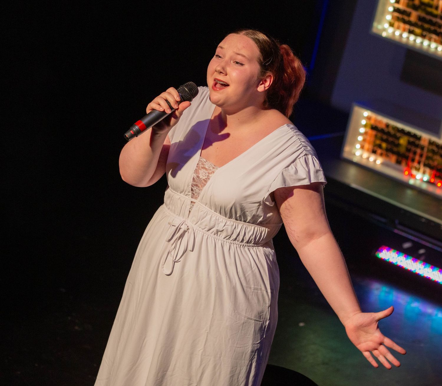 Performer in white dress sings passionately on stage with theatrical lighting effects.