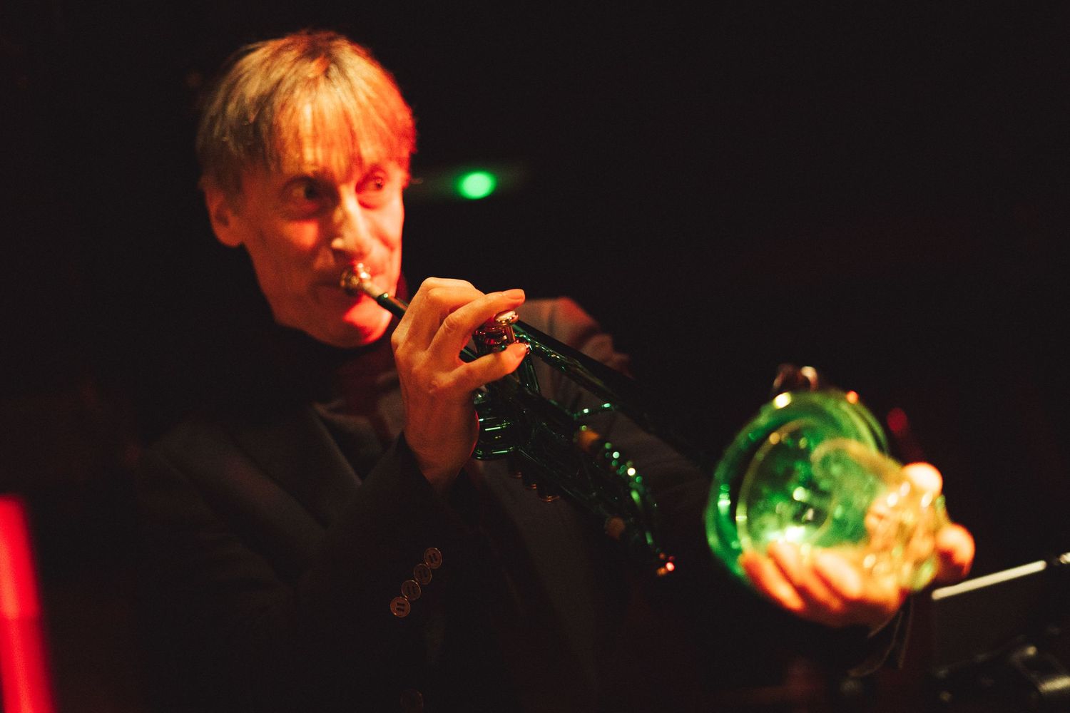 A trumpet player performing with a green glass bottle visible in dramatic red lighting.
