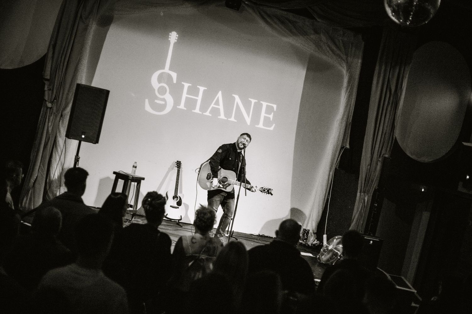 Black and white stage performance with 'SHANE' text projected on the backdrop.