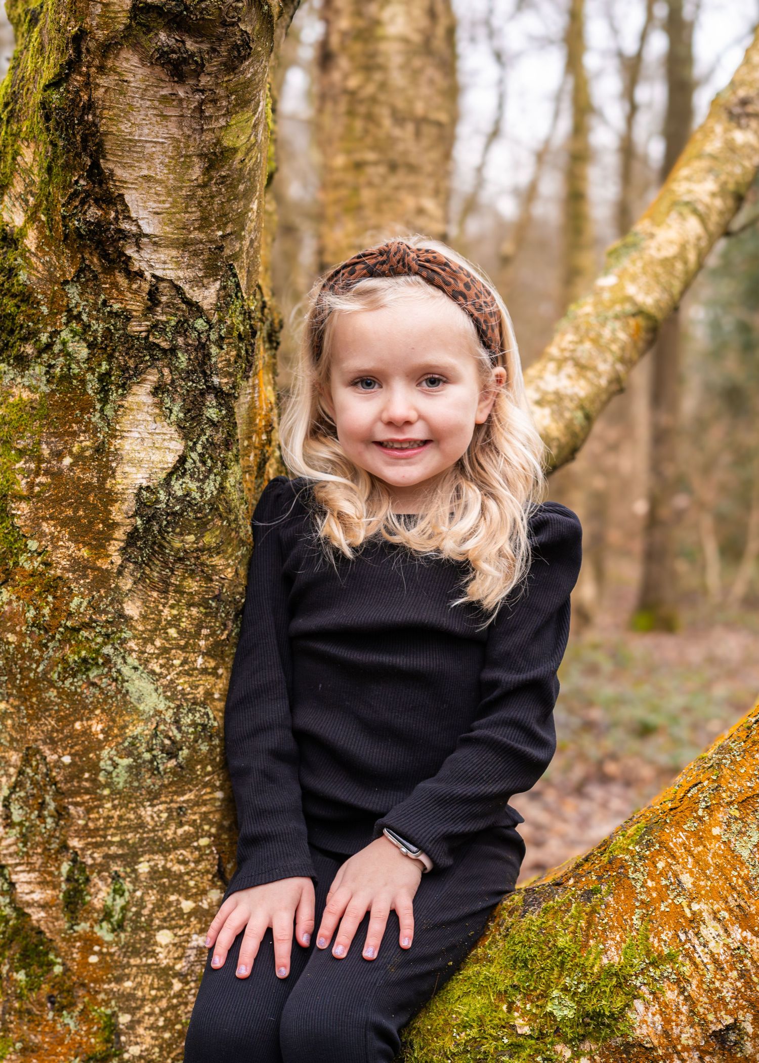 A child in a black sweater sits on a mossy tree branch in an autumn forest setting.