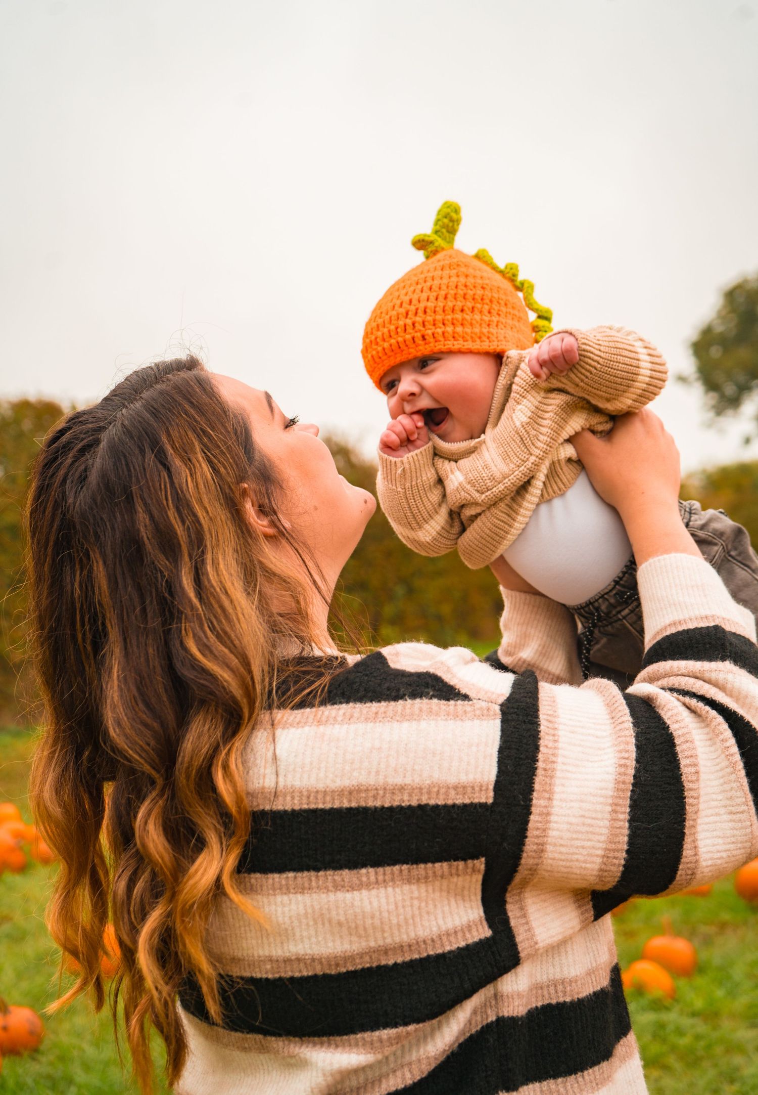 Parent holds baby in striped sweater against backdrop of pumpkin field.
