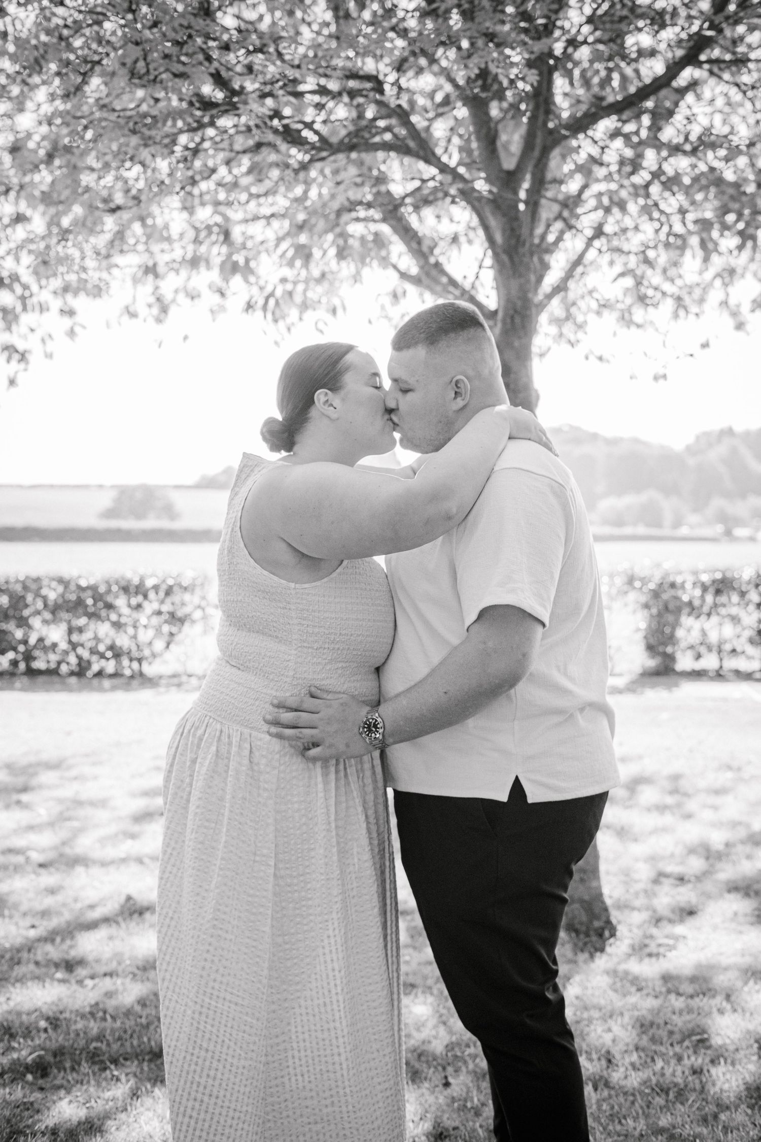 A romantic couple embraces in a park setting with sunlit trees in the background.