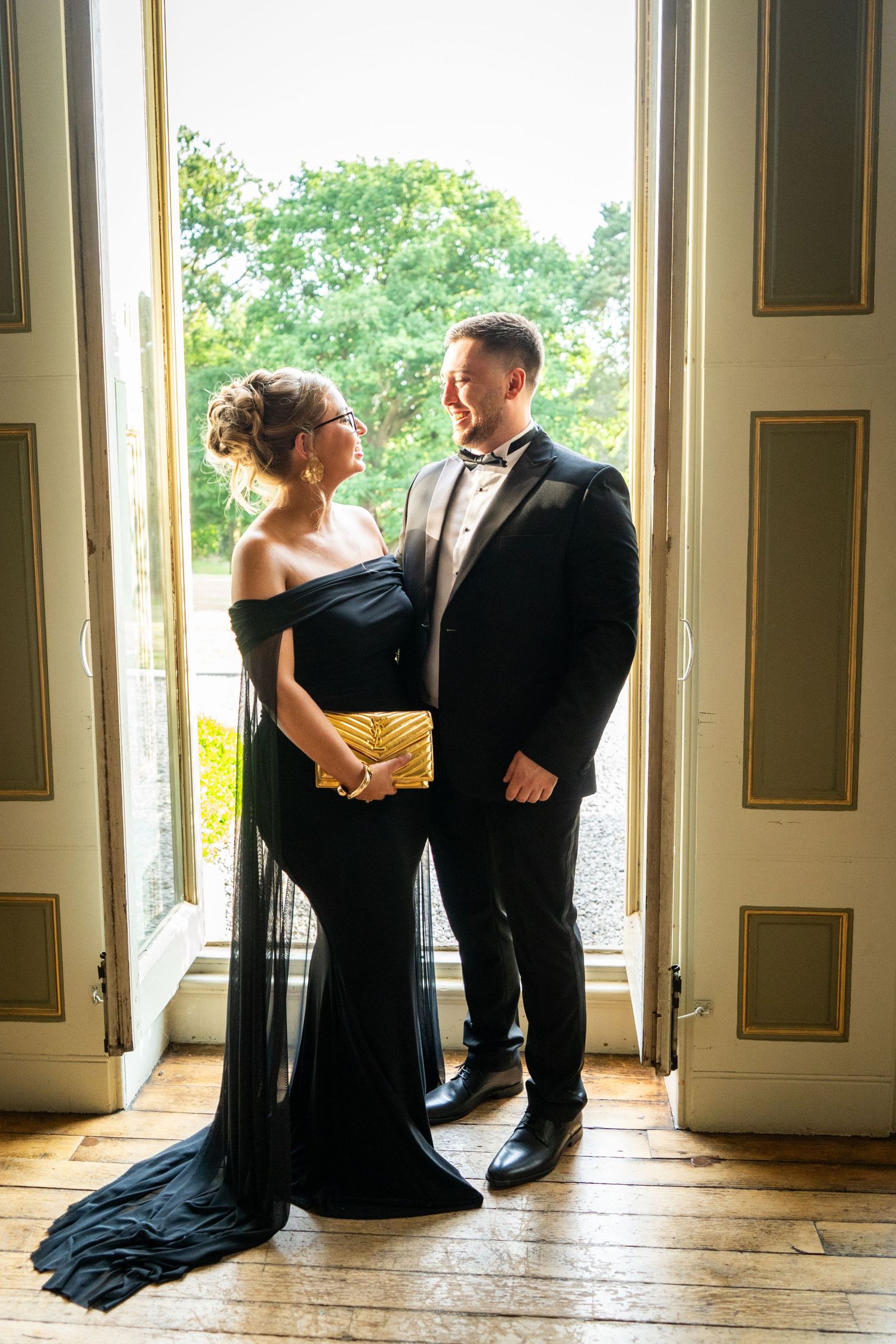 Elegant couple in formal black attire pose in doorway with natural lighting.