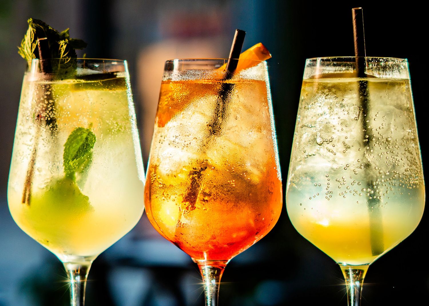 Line of spritz cocktails with varying citrus garnishes on illuminated bar counter.