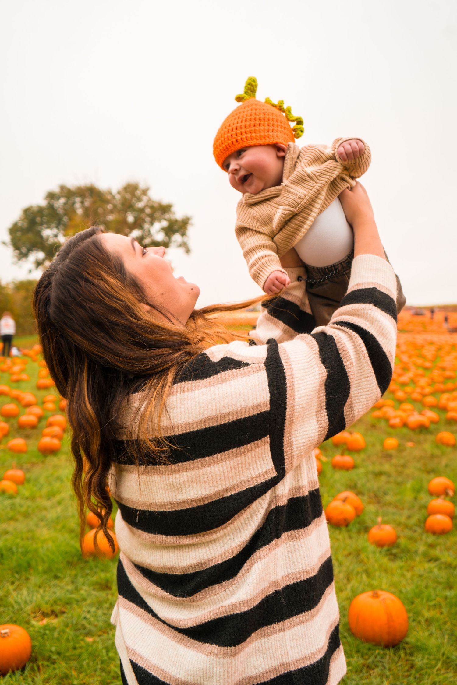 Parent lifts infant wearing orange knit hat in pumpkin patch during fall season.