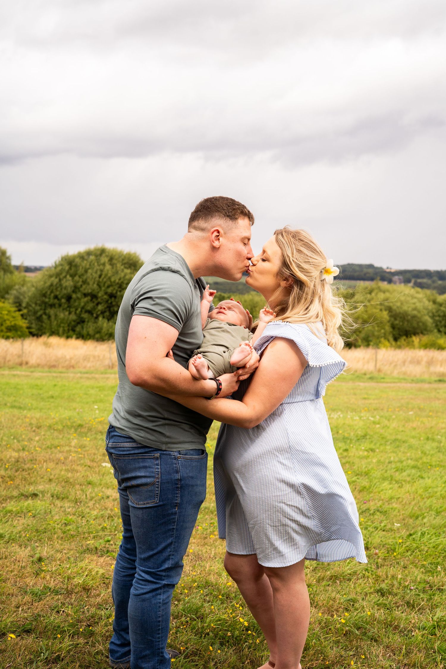 A couple shares a tender moment with their baby in a grassy field under cloudy skies.