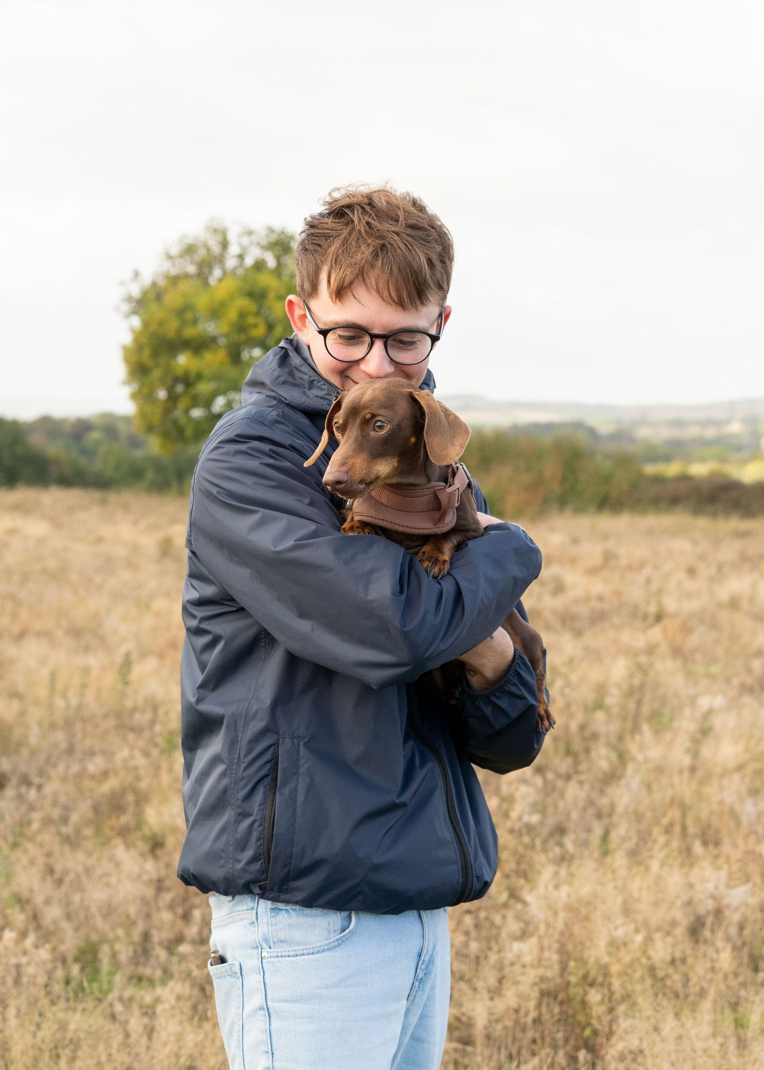 A dachshund puppy being held close and cuddled in a navy jacket in a fall field setting.